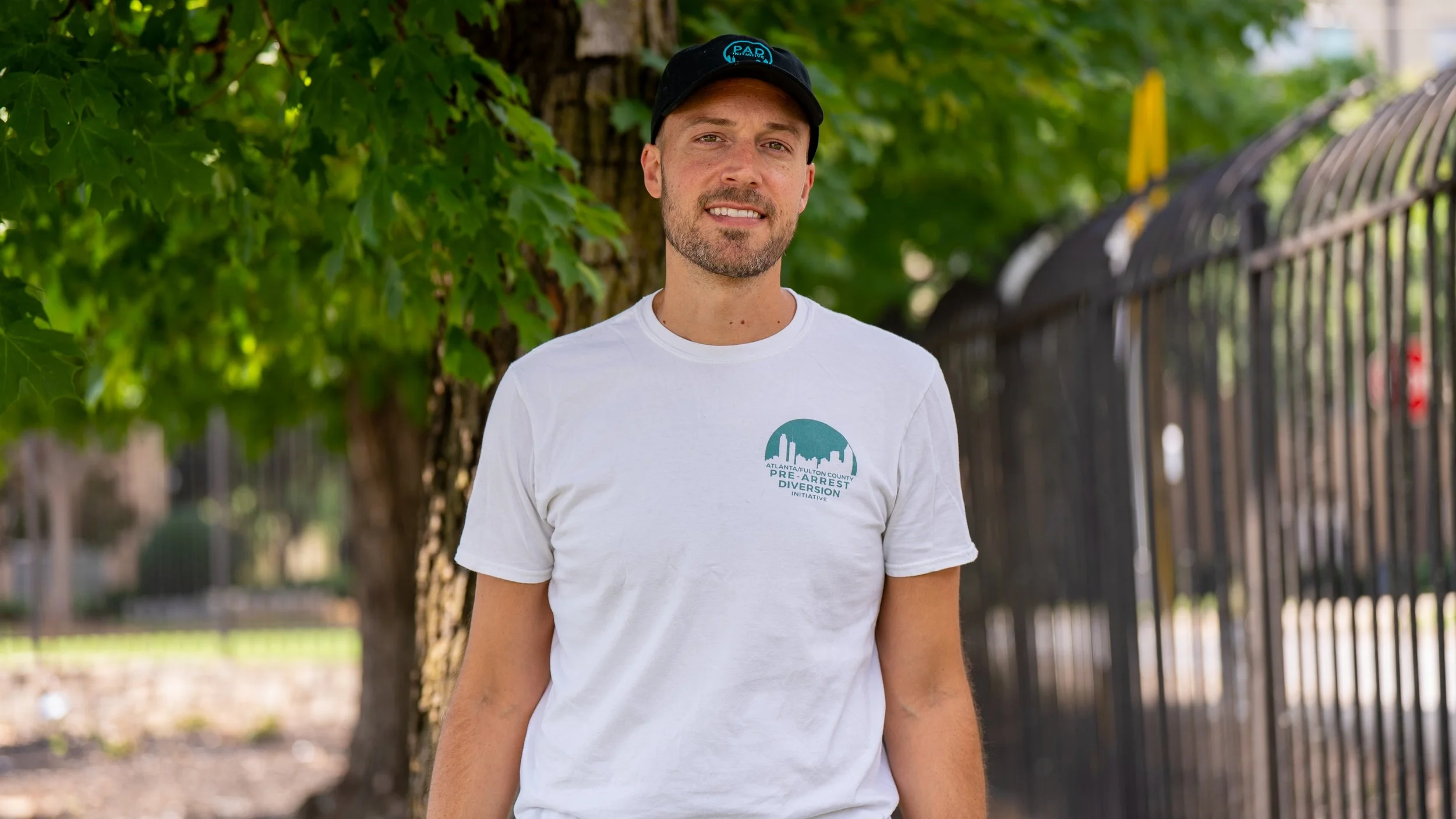  Headhsot of Thaddeus Pettrey, a community Responder at PAD. Thaddeus wears a black PAD hat and a white t-shirt with teal text and graphics that reads:"ATLANTA/FULTON COUNTY PRE-ARREST DIVERSION INITIATIVE".Thaddeus stands in front of a tree.
