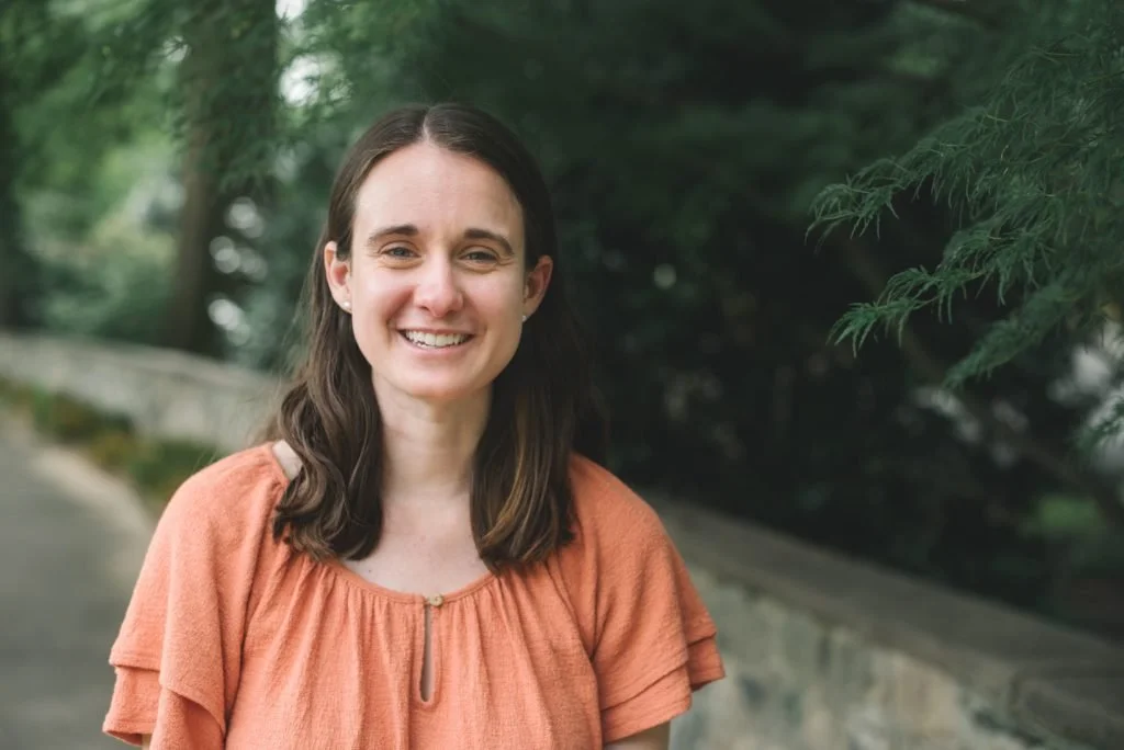 A woman with shoulder-length brown hair smiling outdoors, wearing an orange top, with greenery in the background.