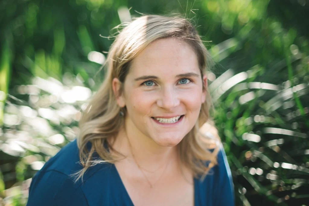 A woman with blonde hair and blue eyes smiling outdoors among green plants.