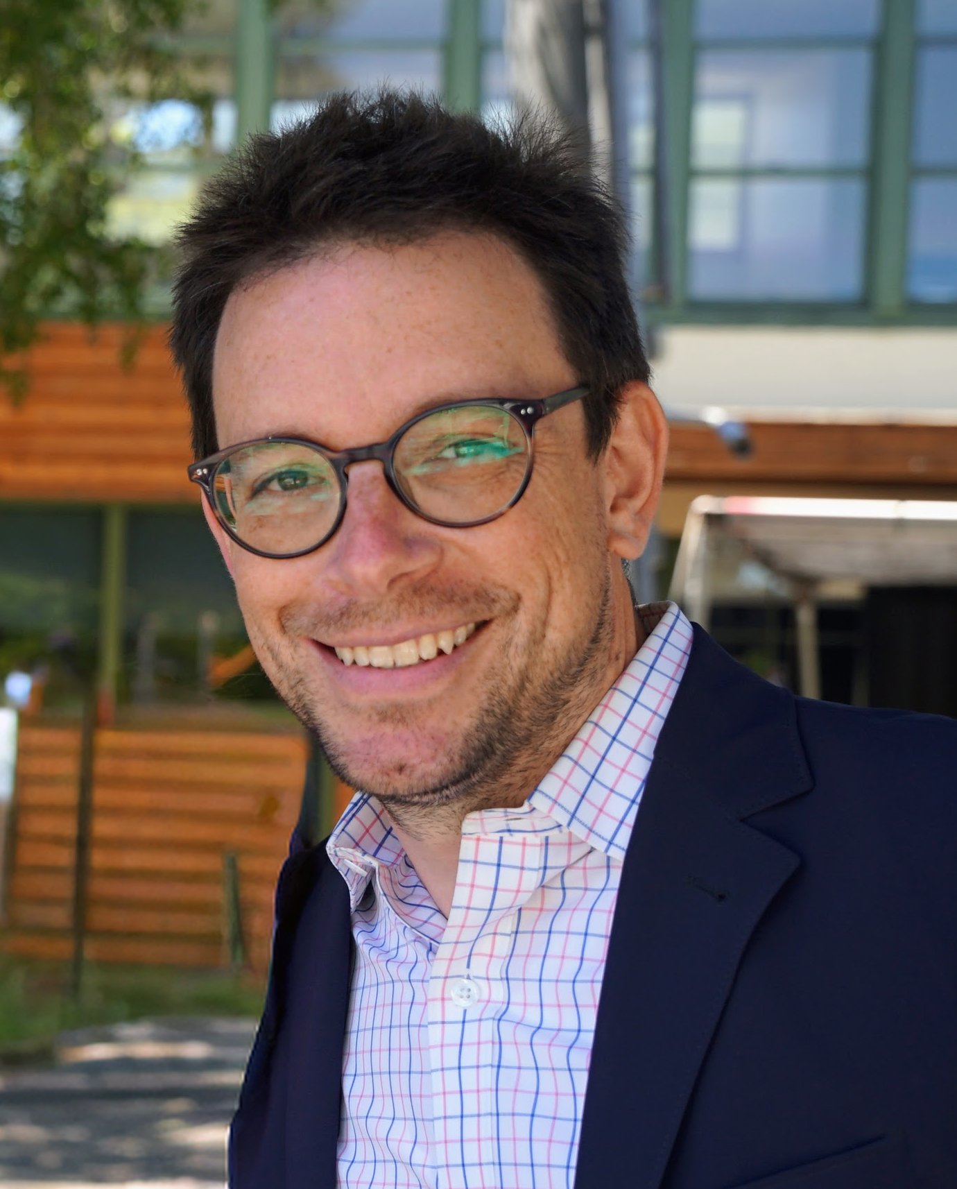 A man smiling, wearing glasses, a navy blazer, and a white checkered shirt with a red and blue pattern. Background includes a modern building with large glass windows and green framing, and wooden fencing.