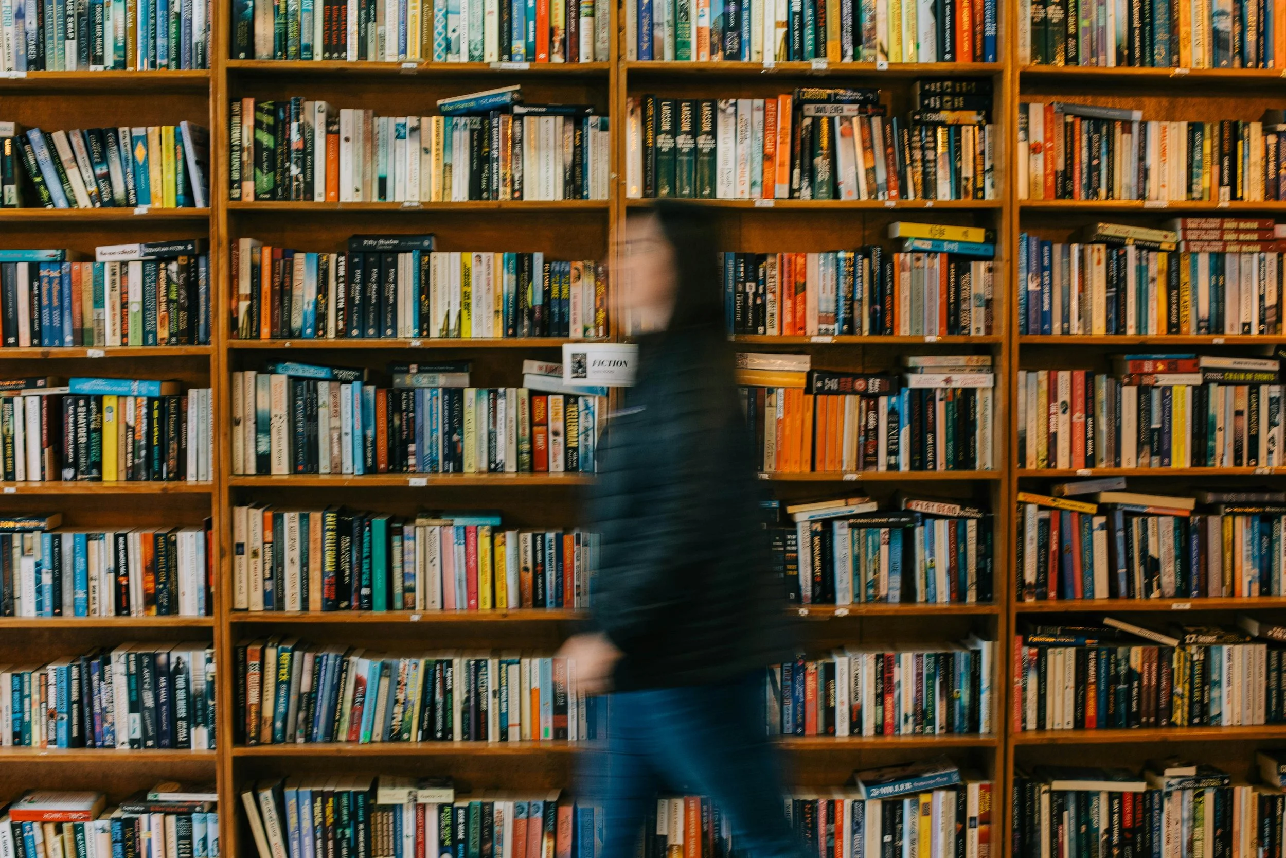 A person walking past wooden bookshelves filled with colorful books in a library or bookstore.