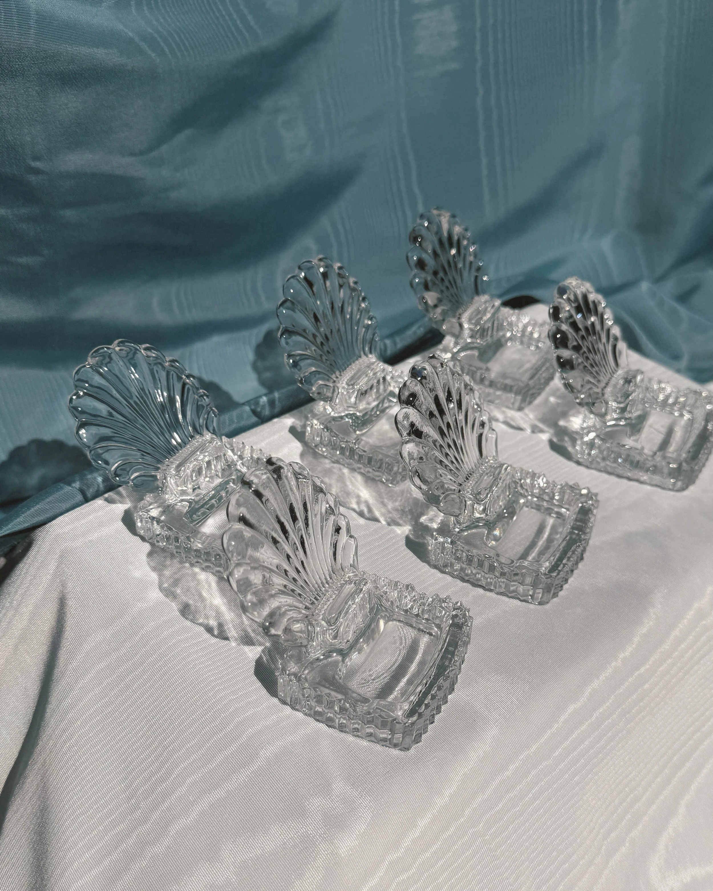 A collection of clear glass or crystal decorative items with a leaf or feather design, placed on a white fabric surface with a light blue fabric background.