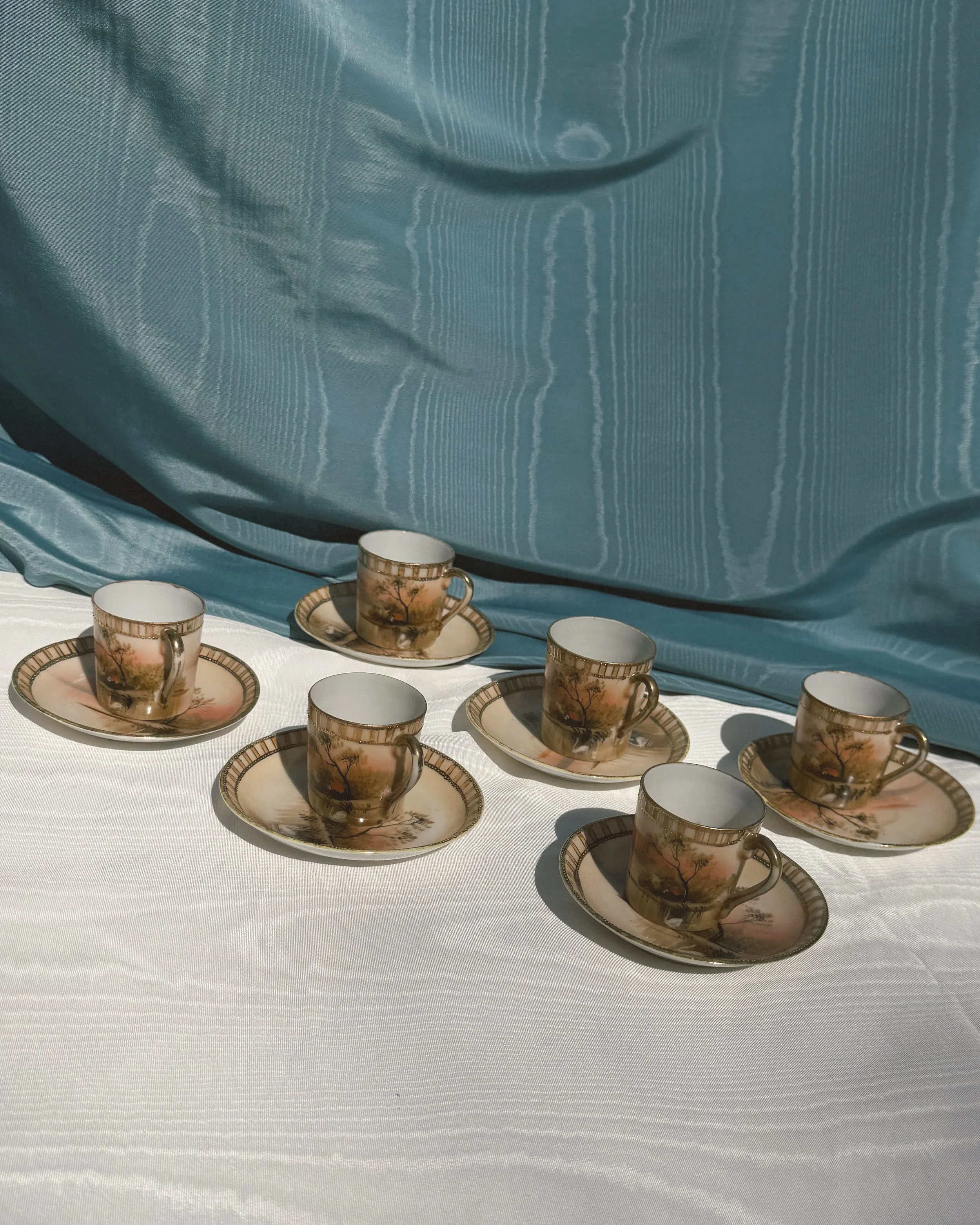 A set of porcelain tea cups and saucers with a landscape design featuring trees and a sunset, arranged on a white tablecloth.