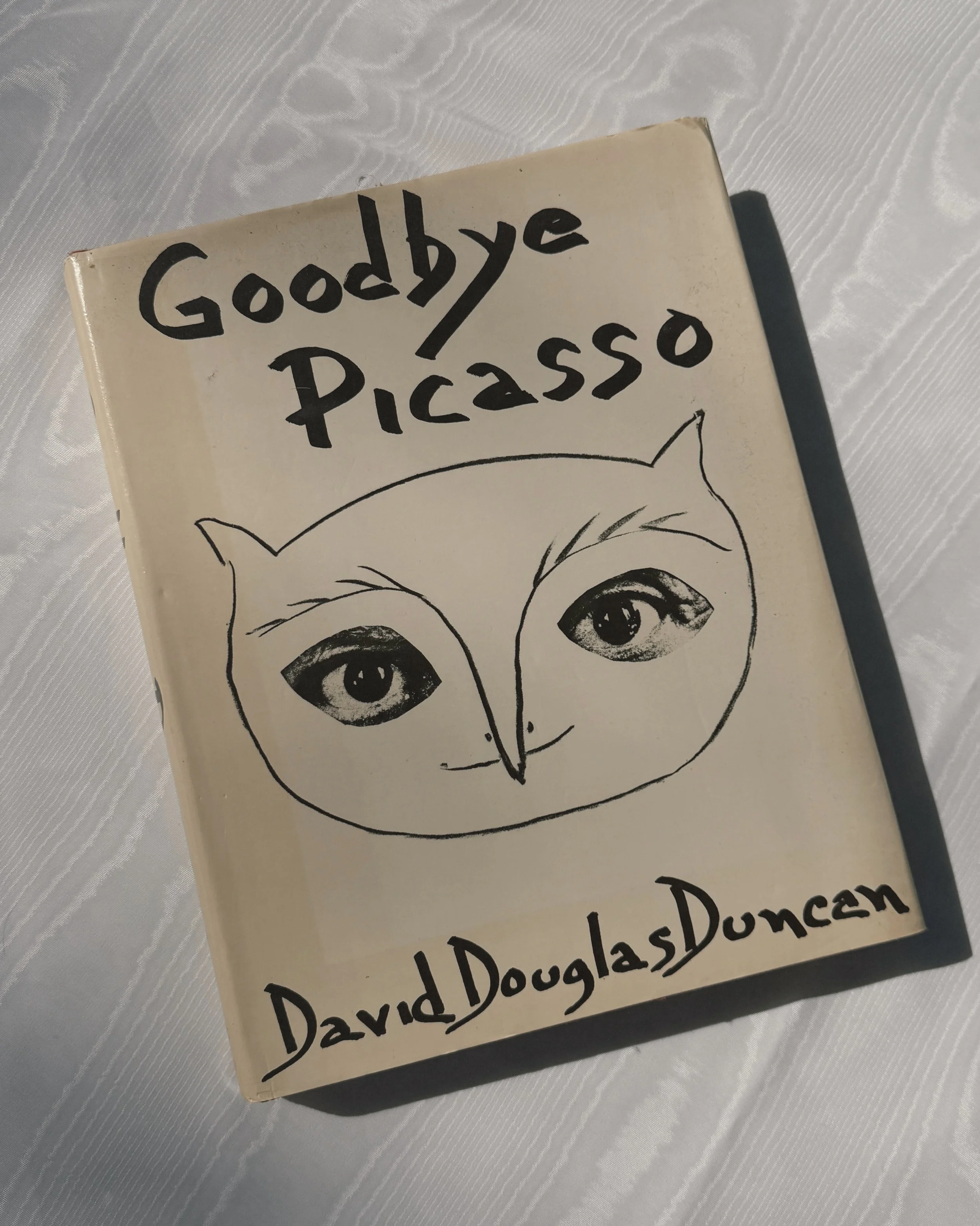 A book titled 'Goodbye Picasso' with a black and white illustration of a face with large eyes on its cover, placed on a shiny silver surface.