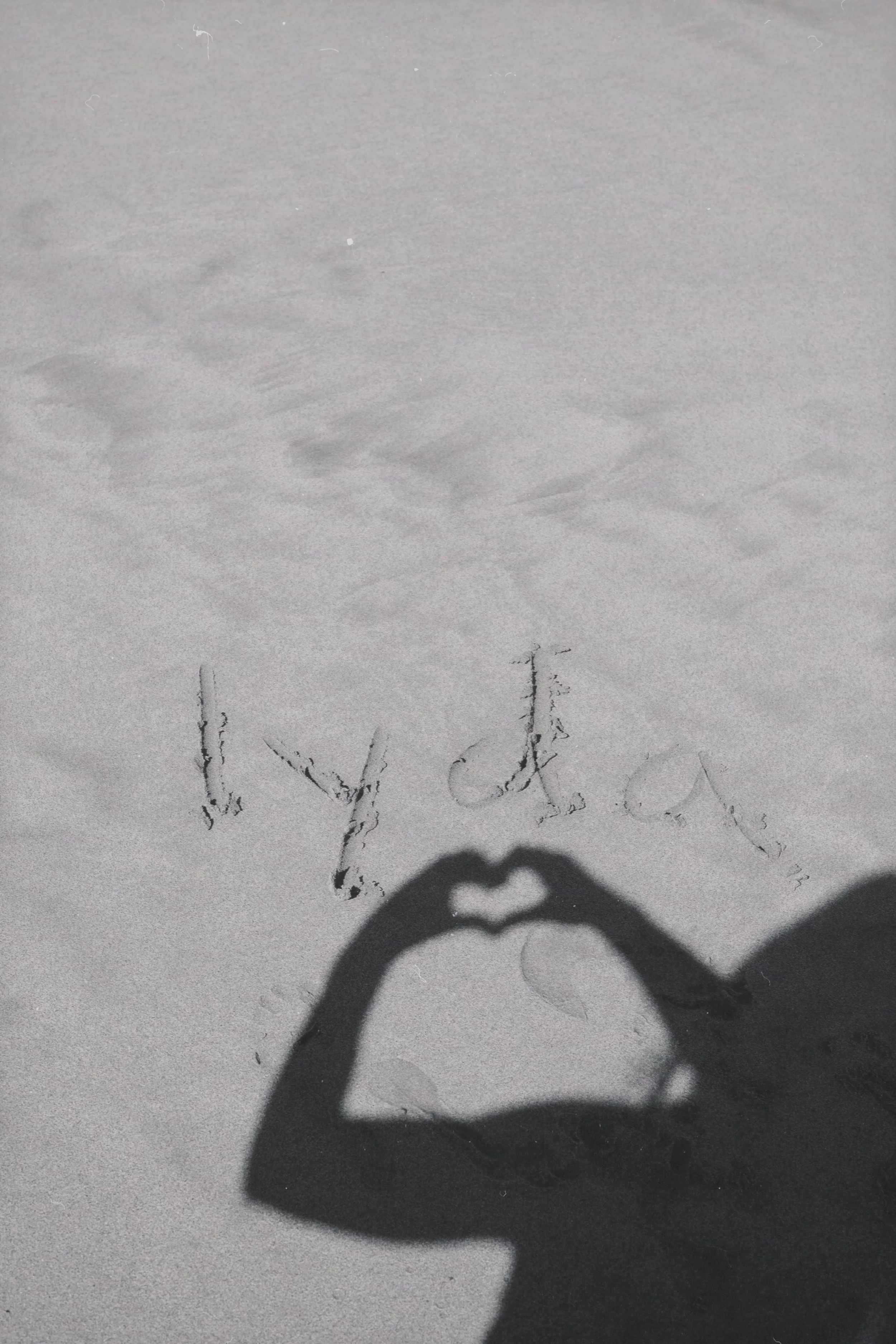 A shadow of a hand making a heart shape is cast onto the snow, with the word 'lyda' written in the sand below