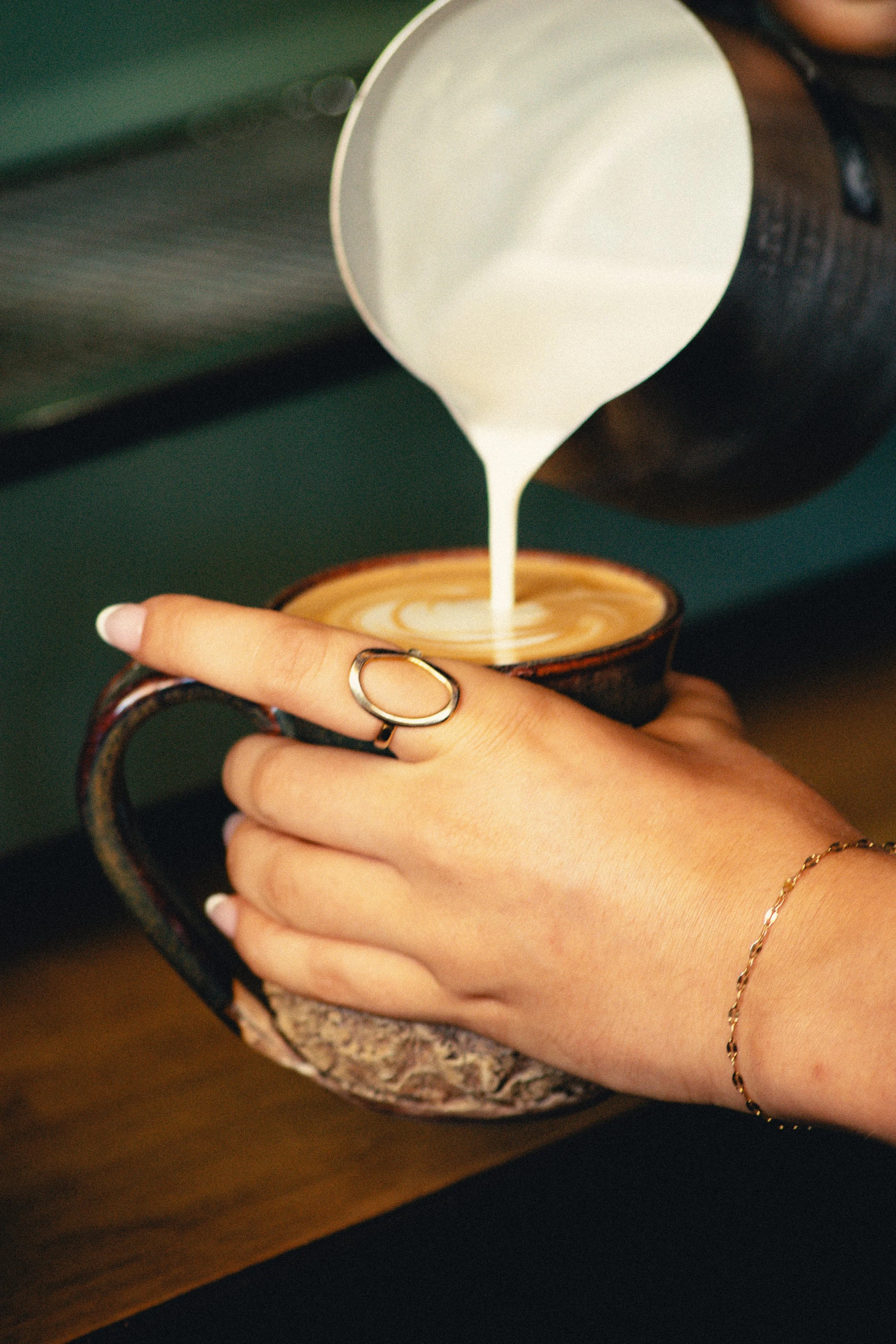 Close-up of a barista pouring milk into a cup of coffee held by a person wearing a ring.