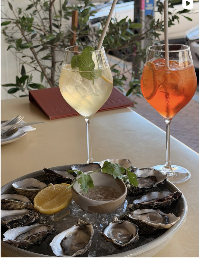 A plate of fresh oysters with lemon slices and dipping sauce, two cocktails—one with mint leaves and the other with orange slices—on a table at an outdoor restaurant.