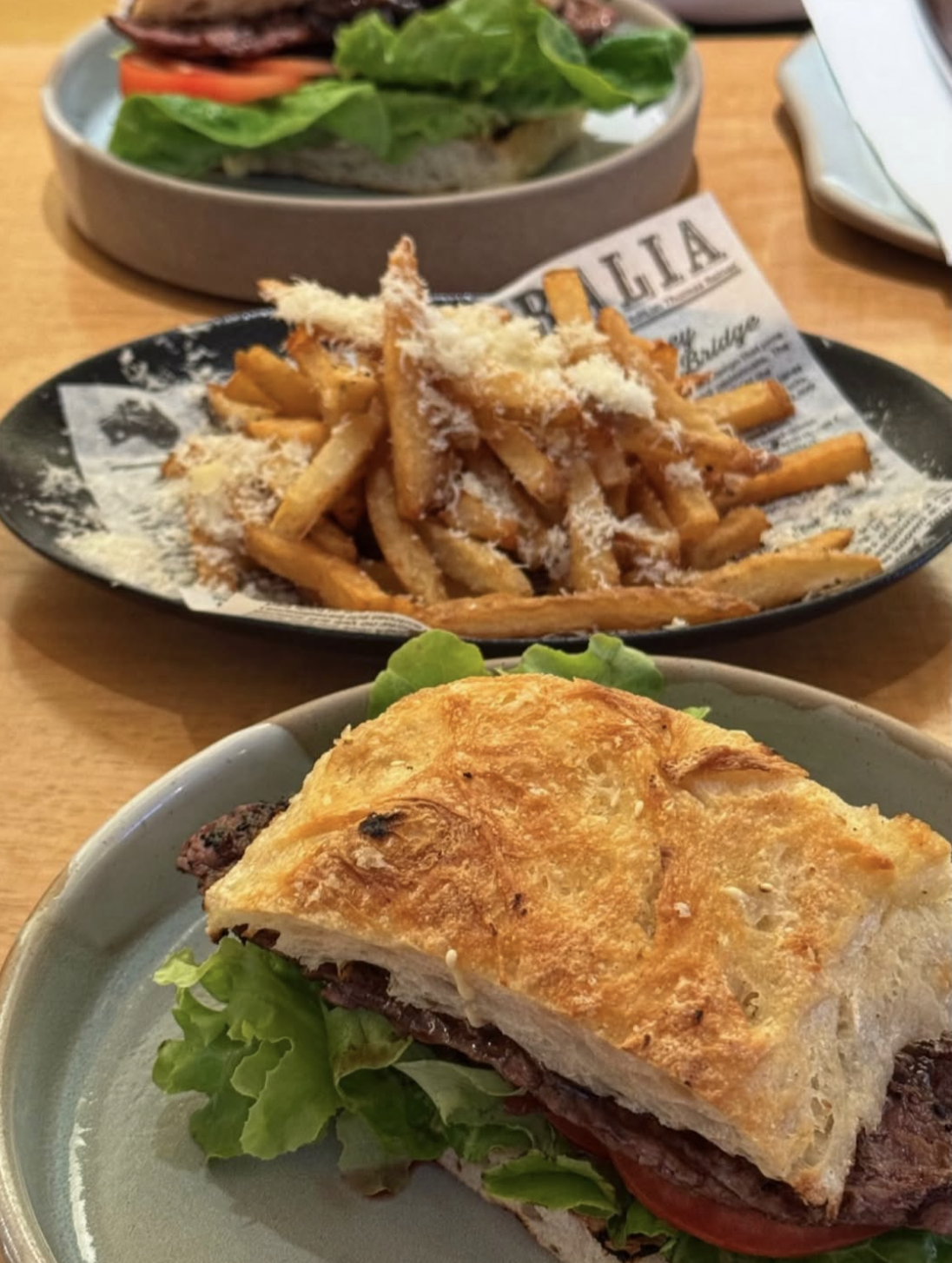 A plate with a slice of baked lasagna containing ground meat, served on a bed of lettuce. In the background, there is a dish of French fries topped with grated cheese. Further back, a sandwich with lettuce, tomato, and possibly meat, is on a separate dish.