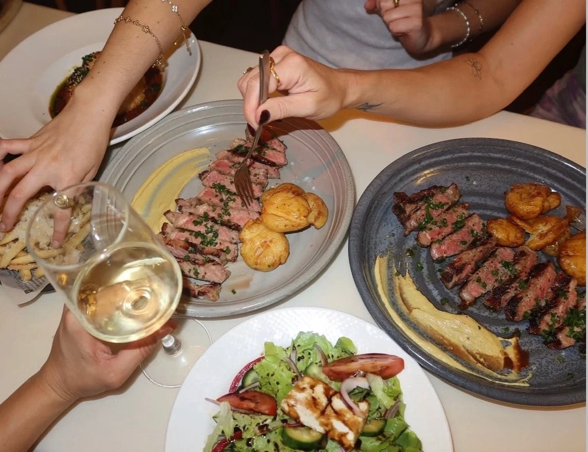 People enjoying a meal with sliced steak, roasted potatoes, a salad, and a glass of white wine.