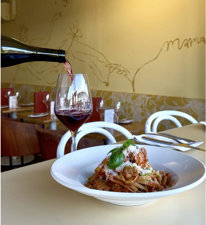 A glass of red wine being poured at a restaurant table with a plate of pasta garnished with basil and cheese, and a background of restaurant seating and wall art.