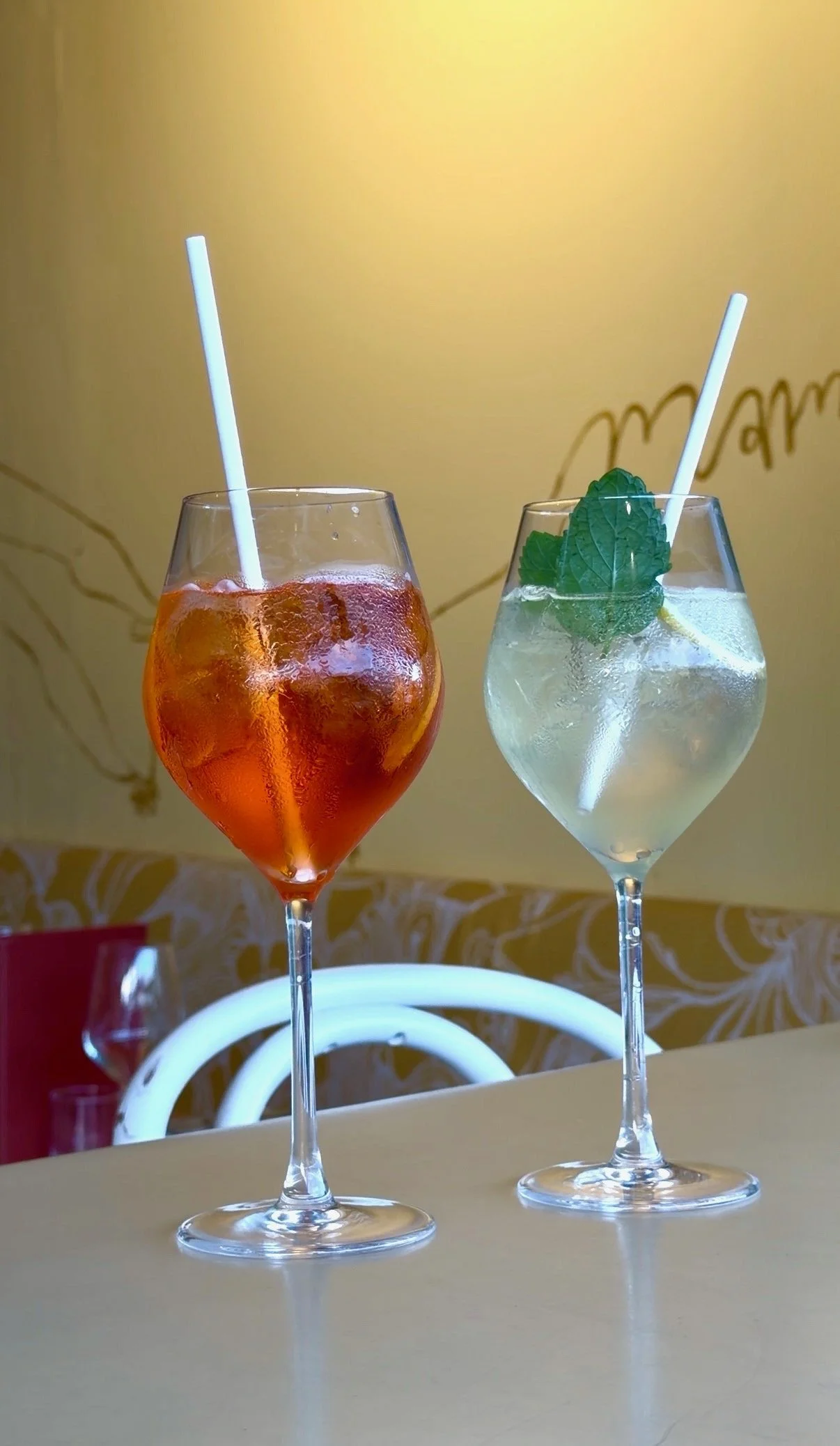 Two cocktails in stemmed glasses with straws, one orange-colored with ice and the other clear with mint leaves and ice, on a table against a yellow wall.