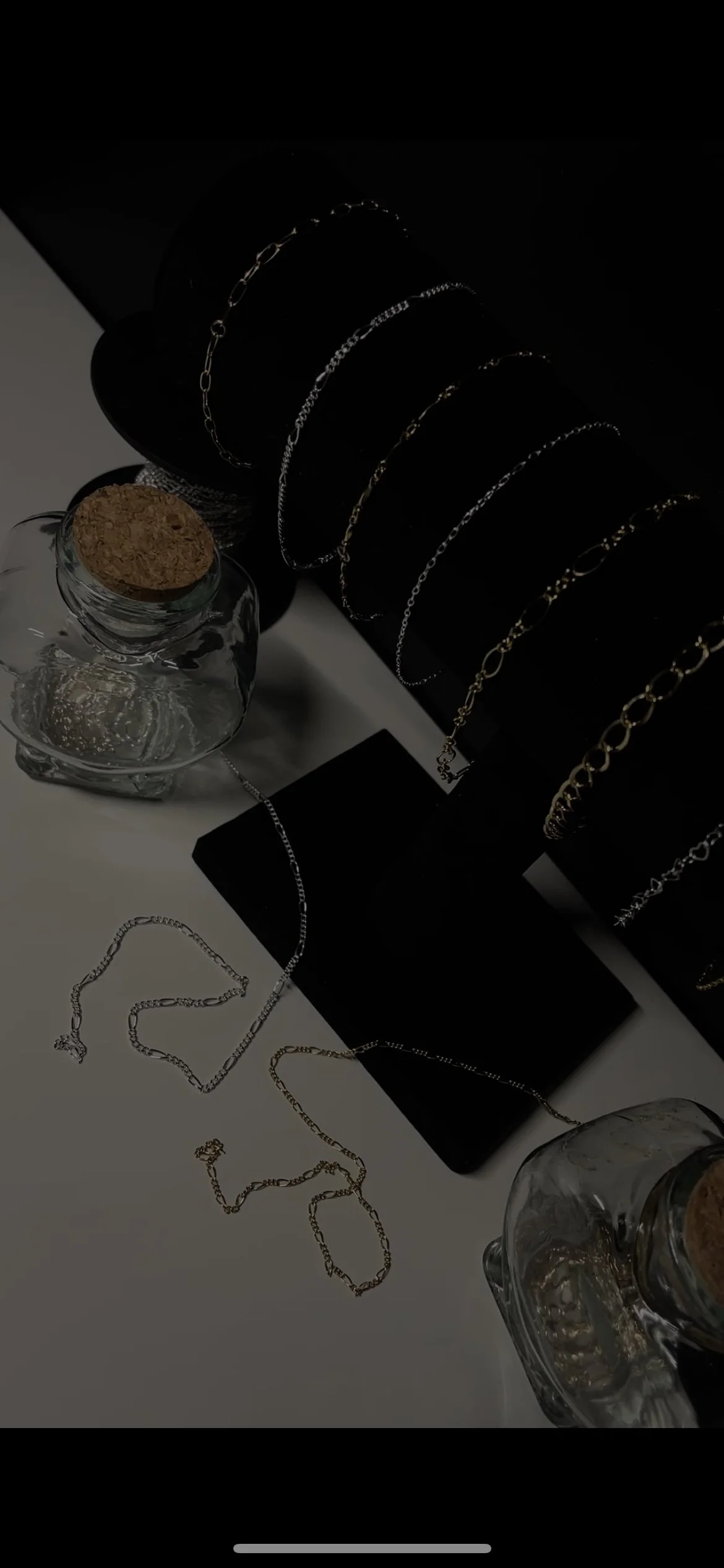 Display of gold and silver necklaces on black cylindrical jewelry stands, with some chains laid out on a white surface, and two glass jars with cork stoppers nearby.