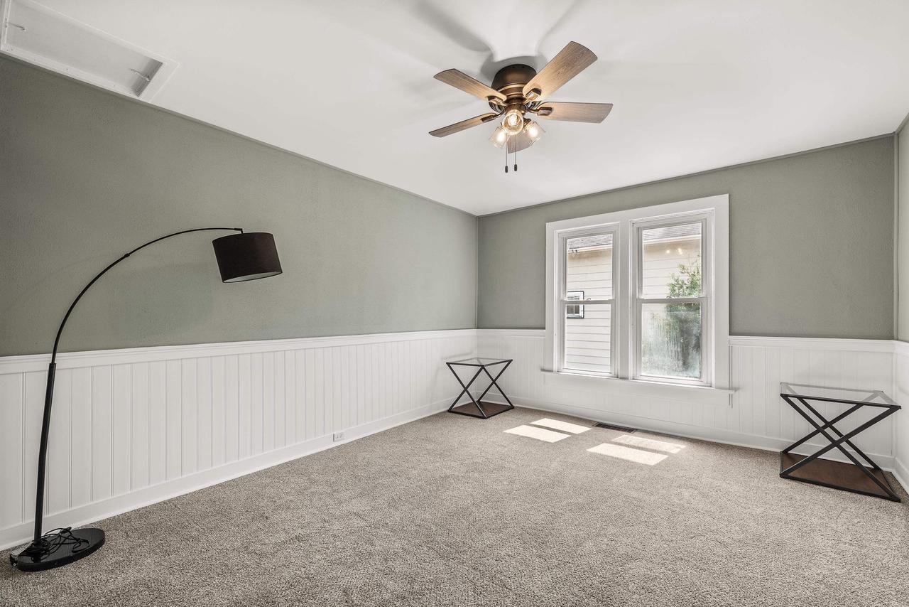 Minimalist room with gray walls and white wainscoting, featuring a ceiling fan, a curved floor lamp, two decorative side tables, and a light gray carpet.