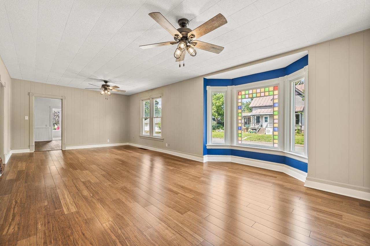 Spacious empty living room with wood flooring, two ceiling fans, and large bay window with colorful trim.