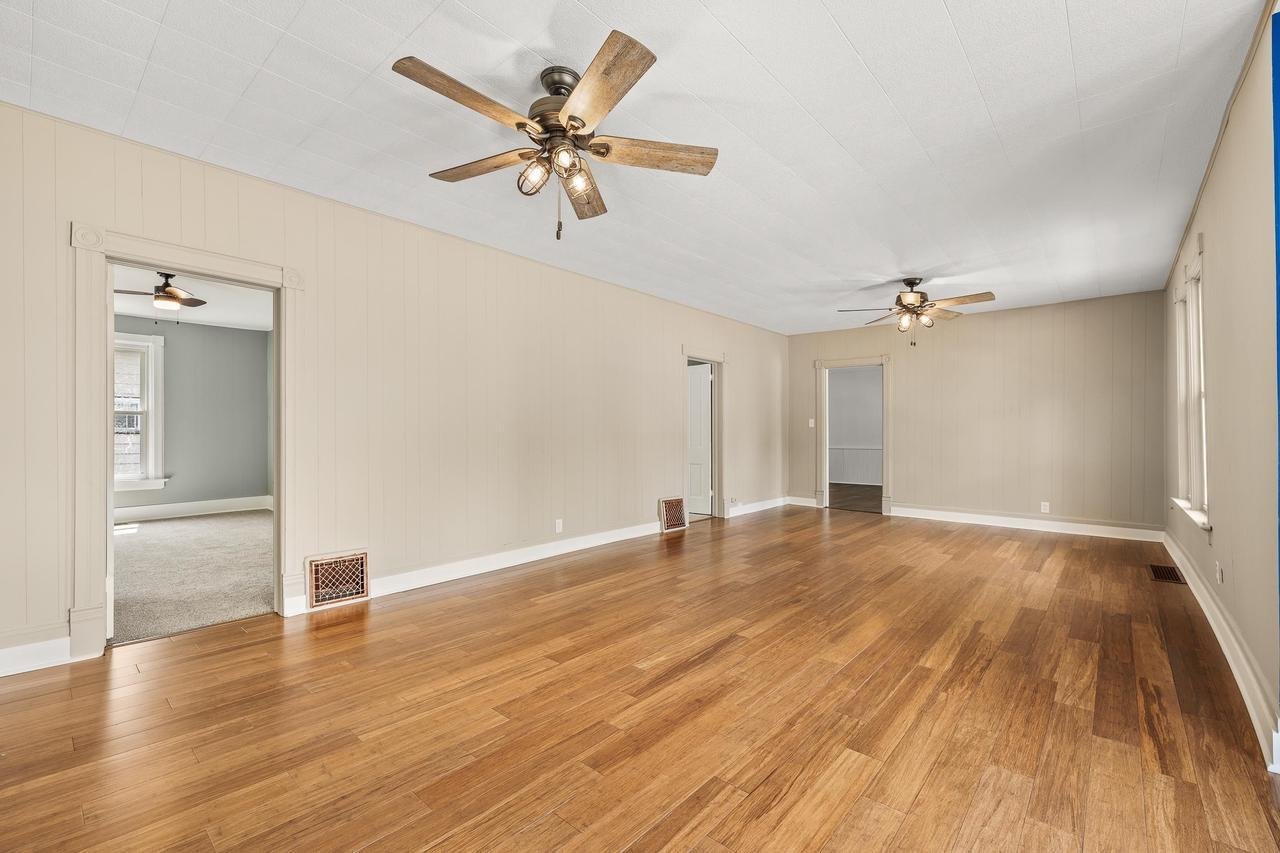 Spacious empty room with hardwood floors, beige walls, white ceiling, two ceiling fans, and multiple doorways, including one leading to a carpeted area.
