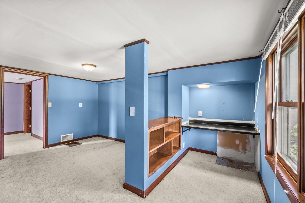 Interior of a room with blue walls, beige carpet, wooden shelves, a small built-in desk, and a window with blinds.