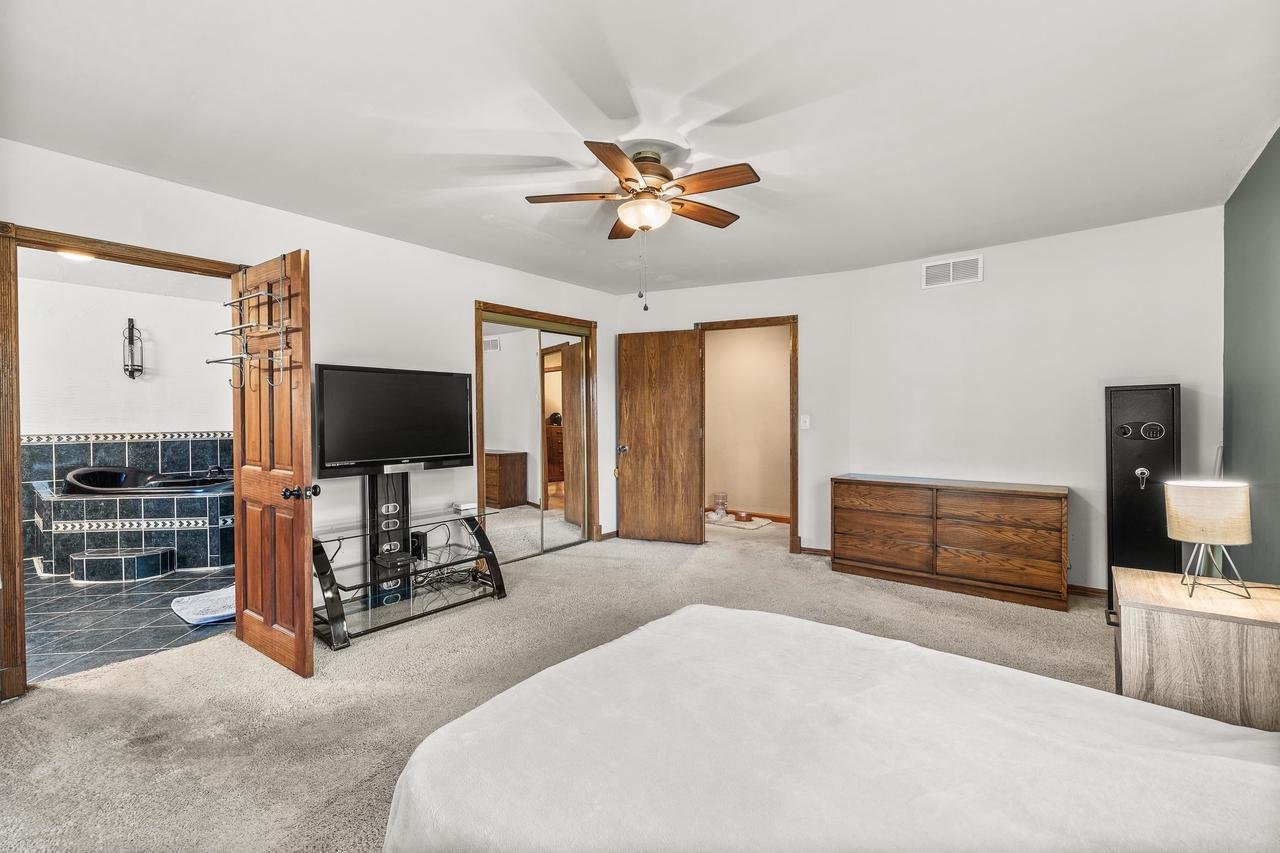Bedroom with carpet, ceiling fan, TV stand, dresser, mirrored closet door, and a safe. Bathroom with a tiled black jacuzzi is visible through an open door.