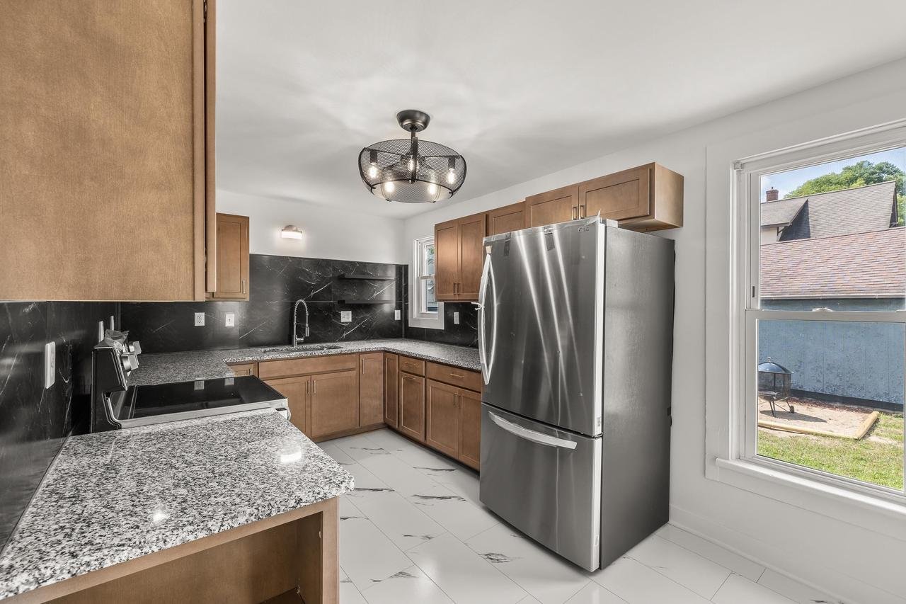 Modern kitchen with stainless steel refrigerator, granite countertops, dark backsplash, wooden cabinets, and tiled floor.