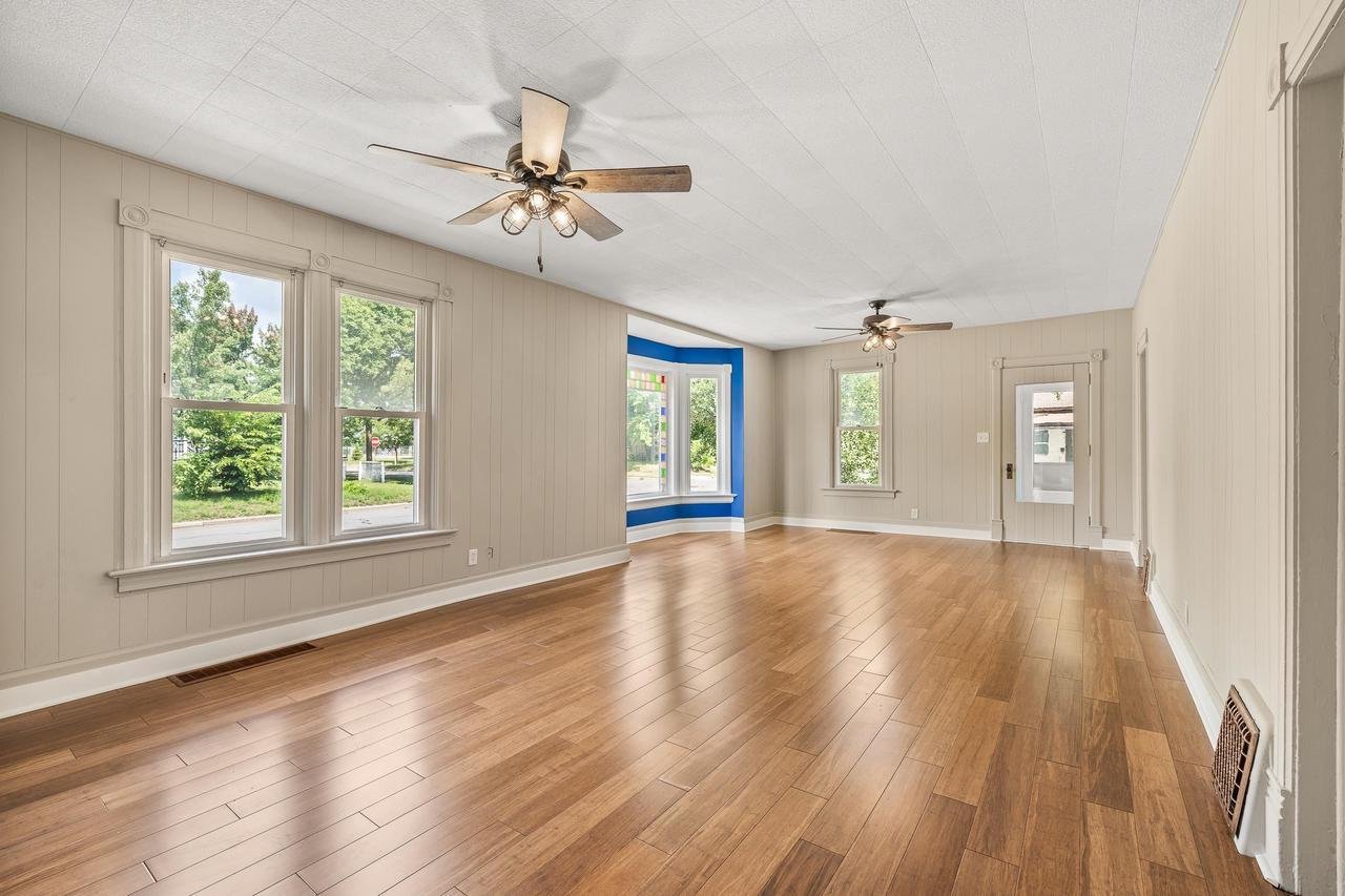 Spacious empty living room with hardwood floors, beige walls, two ceiling fans, large windows, and a door. A bay window with a blue accent is visible in the middle.