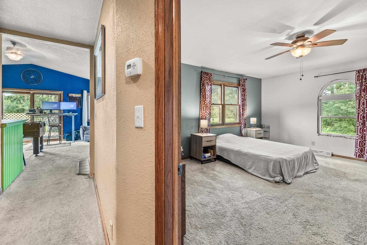 Interior view of a house featuring a bedroom and an adjacent room. The bedroom has a bed with a simple cover, side tables, and a ceiling fan with wooden blades. It includes a window with patterned red curtains. The adjacent room has blue walls, dual