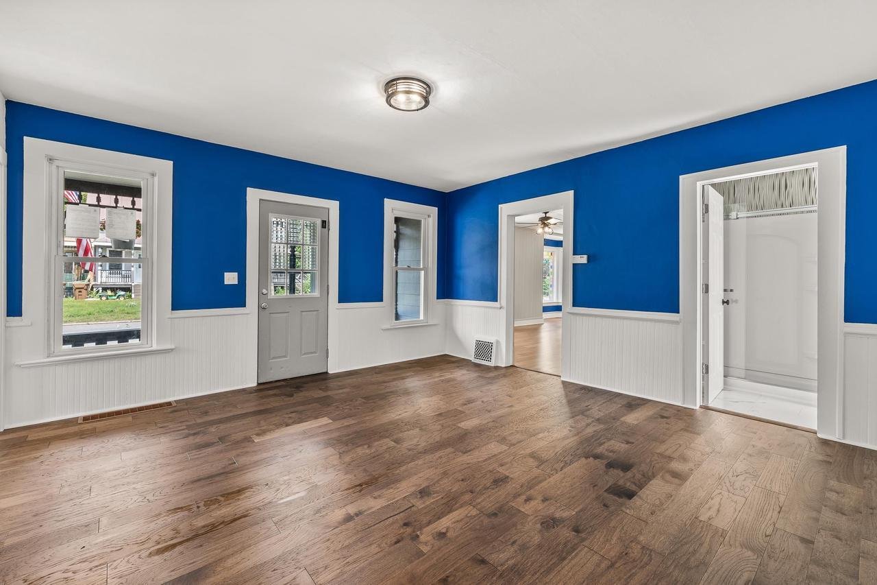 Bright room with blue walls, white trim, wooden floors, and a ceiling light. Includes windows, a wooden door, and a visible doorway to another room.