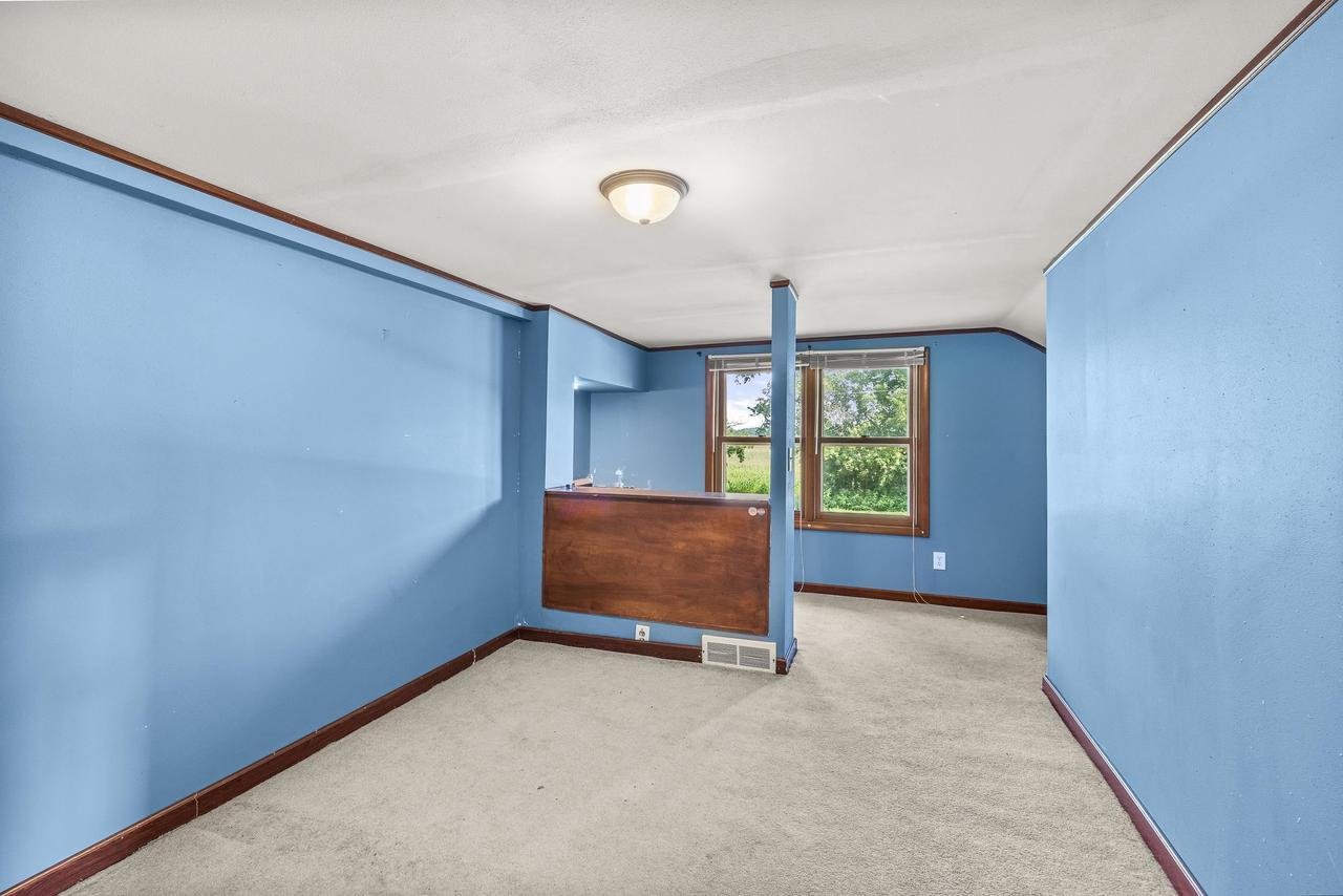 Empty room with blue walls, beige carpet, and a ceiling light. The room features a window with a view of greenery outside and a wood-paneled section under the window.