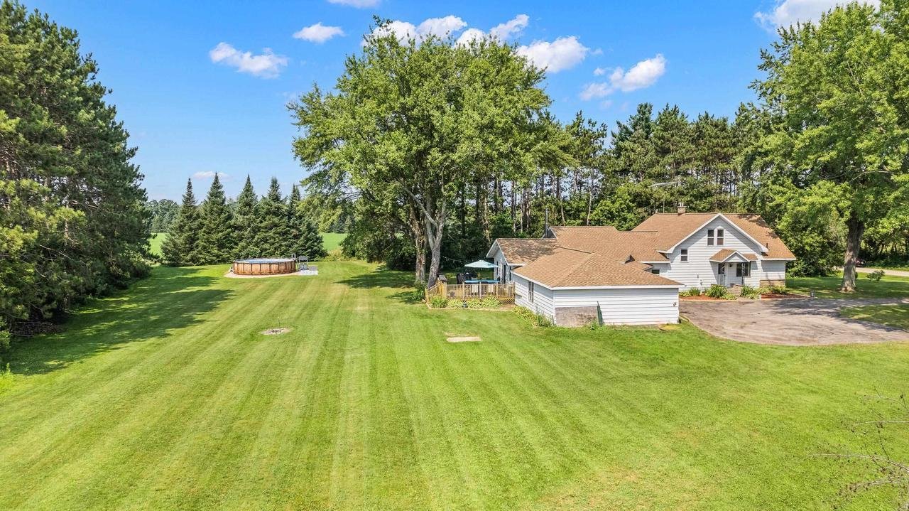A suburban residential property featuring a white house with a brown roof, surrounded by a spacious, well-manicured lawn. The backyard includes an above-ground pool and is bordered by tall trees, creating a private, scenic environment. A large tree s