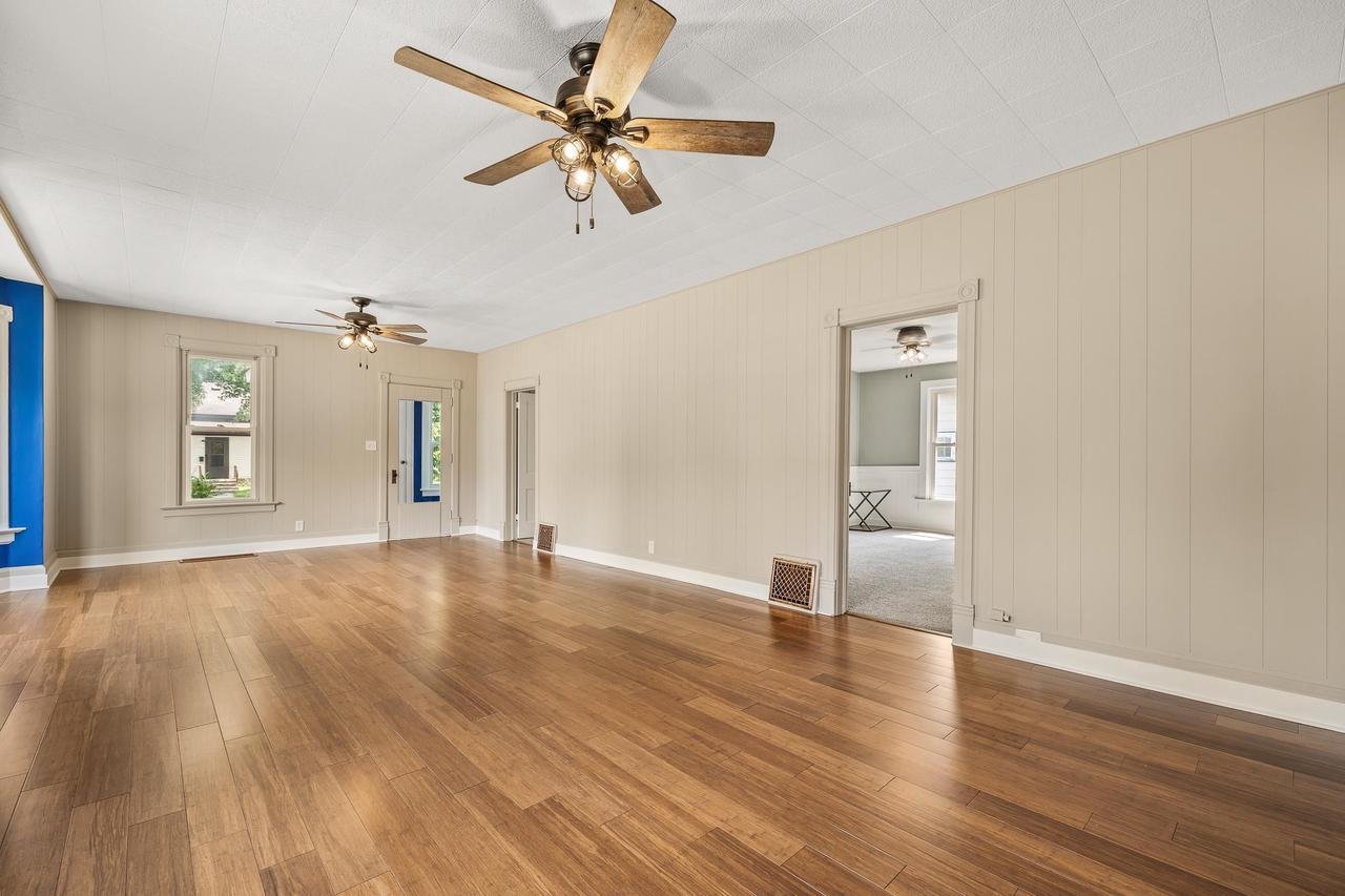 Spacious empty room with wooden floors, beige walls, and a white ceiling featuring two ceiling fans. The room has large windows letting in natural light and visible vents on the walls. A doorway leads to an adjacent room with carpeted flooring.