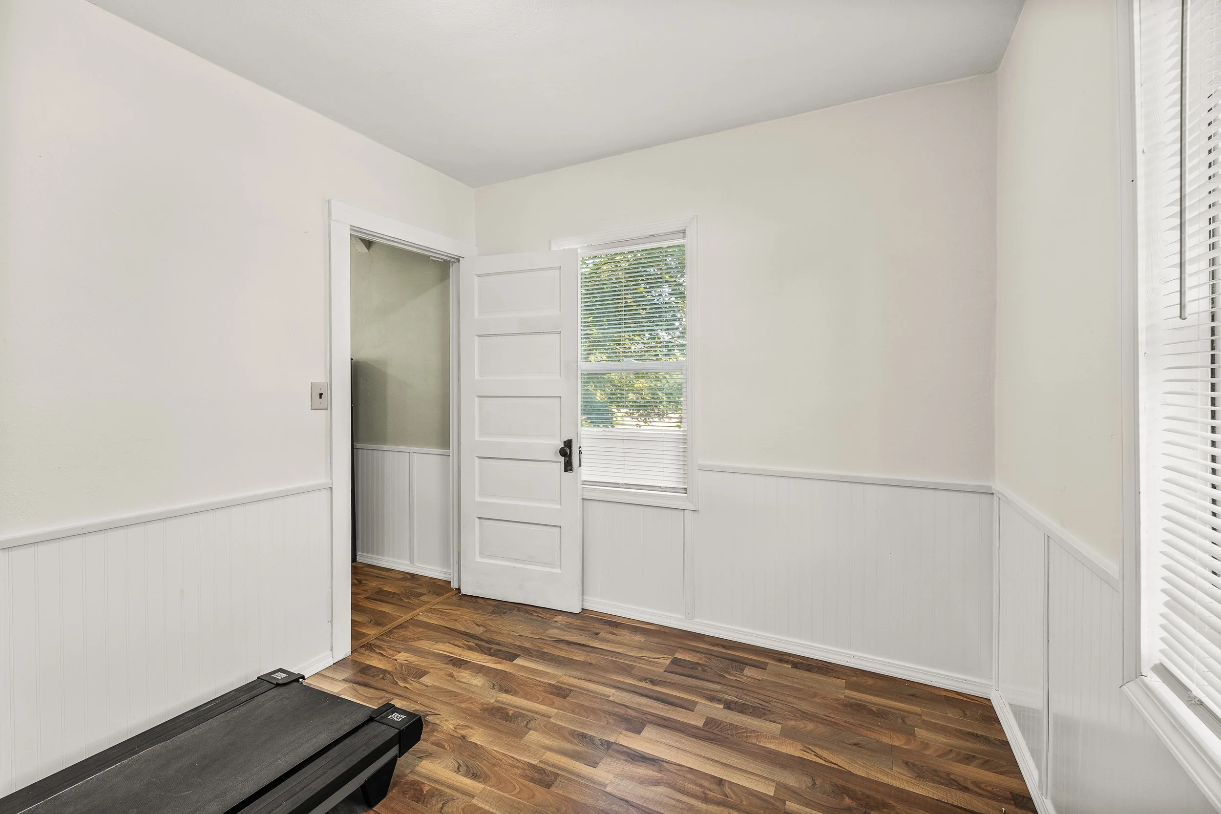 Empty room with open door, wood flooring, white walls, and window blinds.
