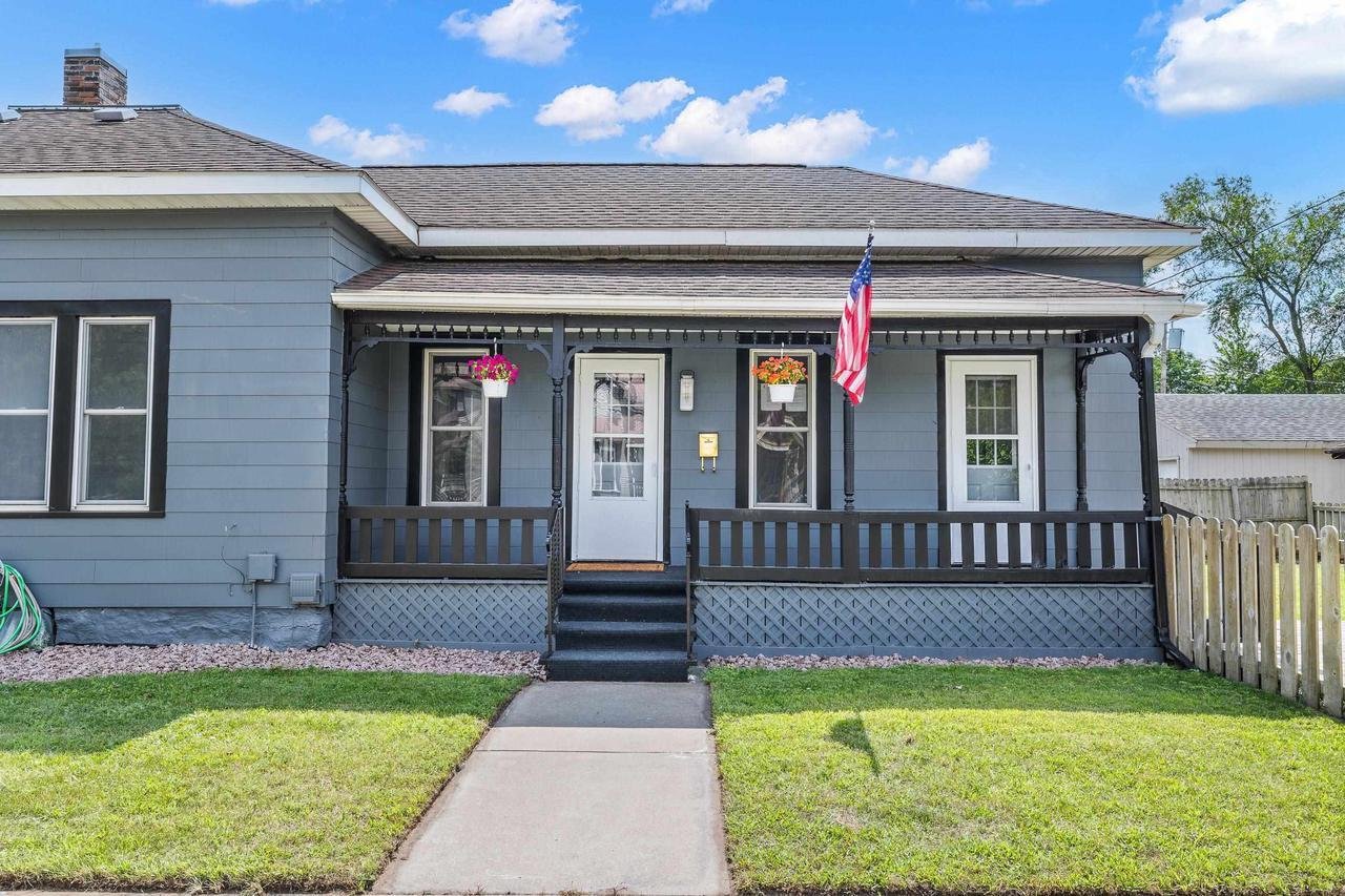 Gray single-story house with a porch, American flag, hanging flower baskets, and a small lawn.