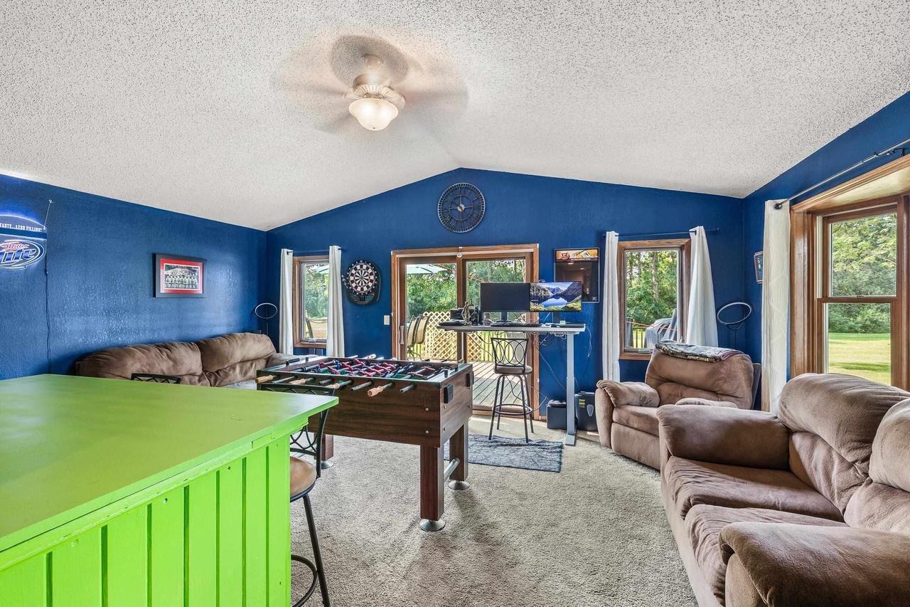 Recreation room with blue walls, featuring a foosball table, green bar counter, brown sofas, a dartboard, ceiling fan, and large windows with white curtains opening to a green lawn.