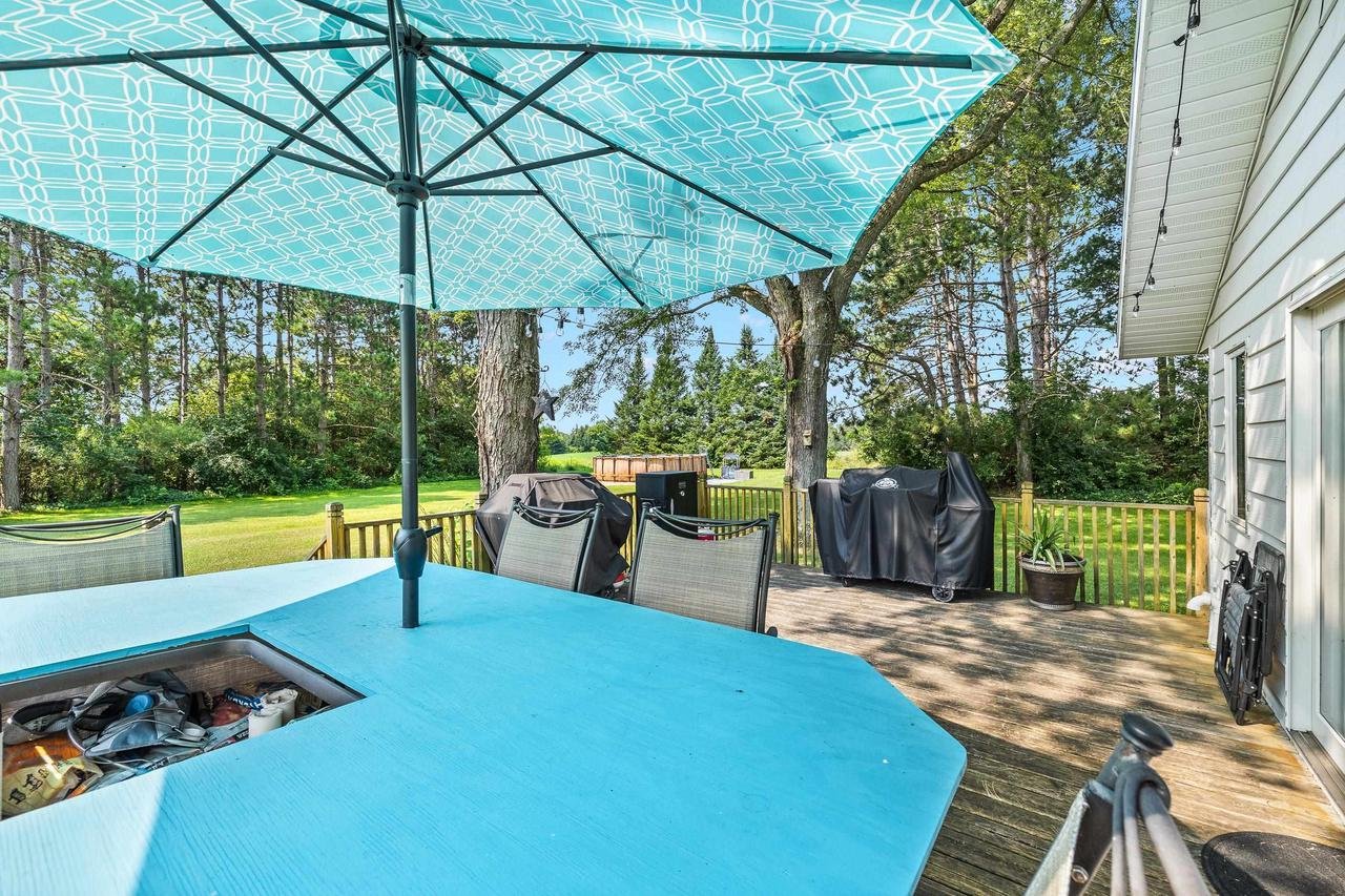 Outdoor patio with blue umbrella, wooden deck, and lawn chairs; grill covered with black tarp; lush green trees and grass in the background.