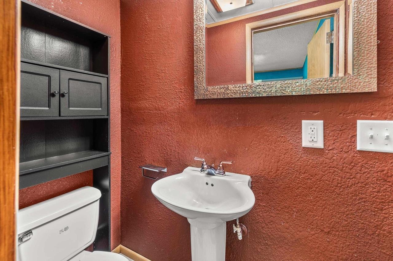Small bathroom with red textured walls, white pedestal sink, toilet, large mirror, black cabinet above toilet, and light switch.