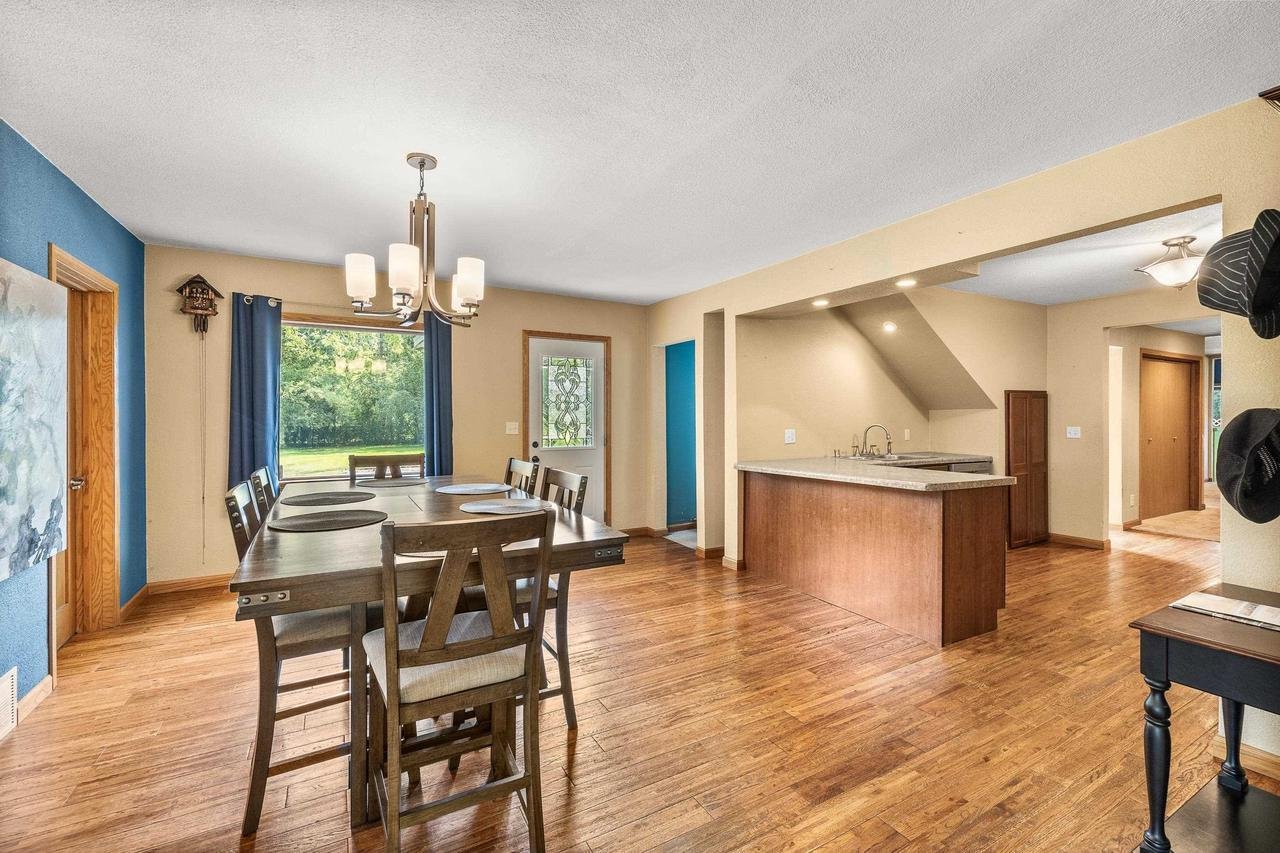 Open-concept dining and kitchen area with wooden flooring, a large dining table with chairs, modern chandelier, blue curtains, and a kitchen island with a sink.