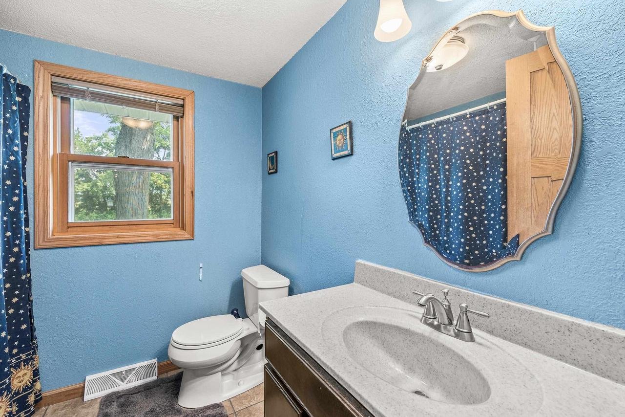 Bathroom with blue walls and decor, featuring a white toilet, oval mirror, sink with faucet, and a window with a wooden frame.