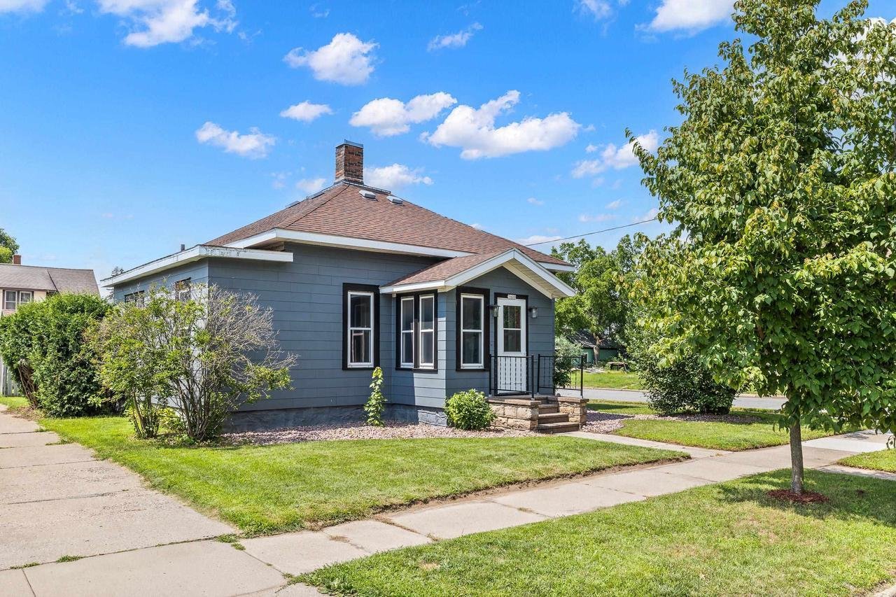 A small blue house with a red roof and a chimney. The house has a porch with steps leading up to it and is surrounded by a green lawn with a few trees and shrubs. A sidewalk runs in front of the house under a partly cloudy blue sky.