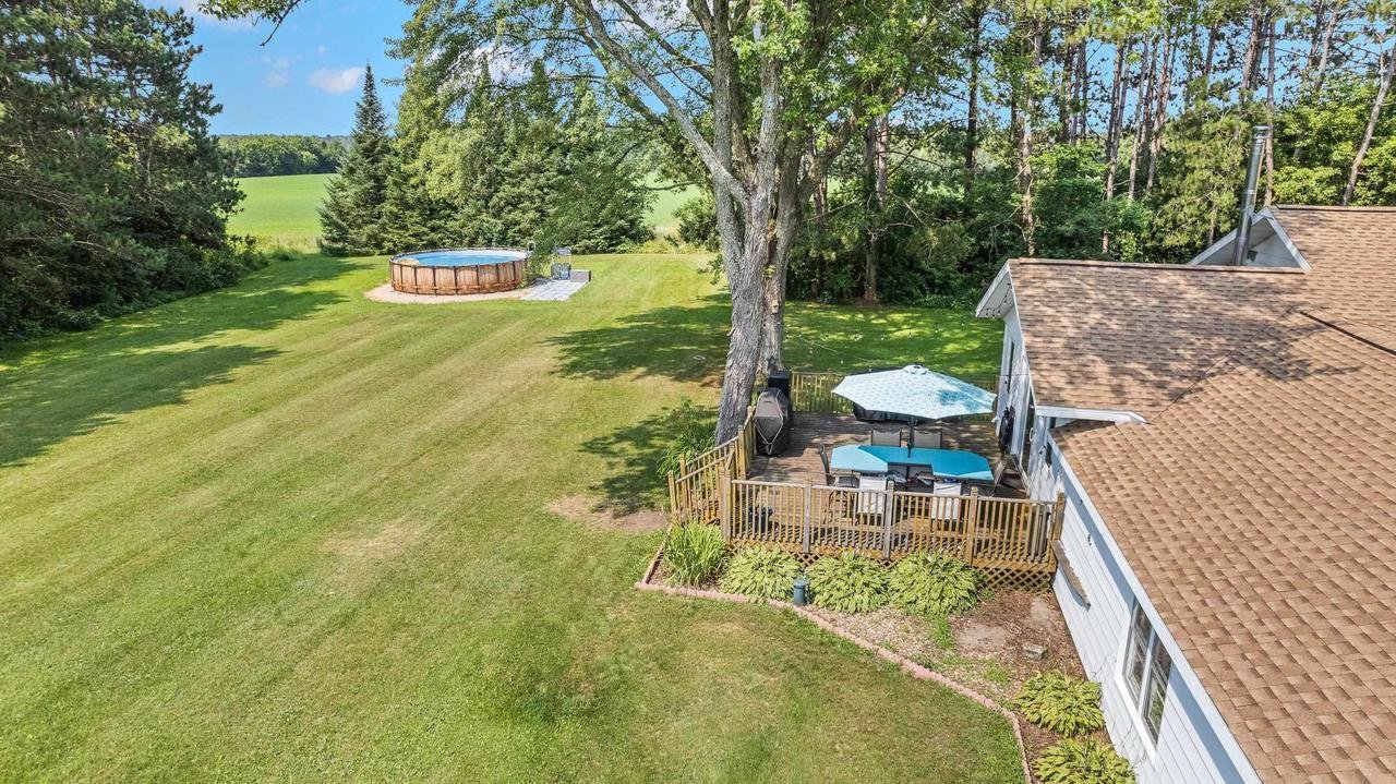 Aerial view of a backyard with an above-ground pool and a patio. The pool is surrounded by green grass and trees. The patio, attached to a house, features outdoor furniture and an umbrella. A large tree provides shade to the patio area.