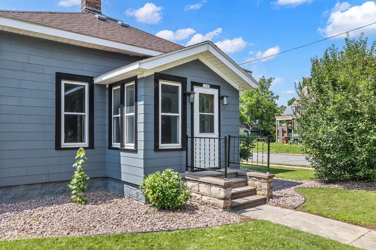 Blue house with a front porch, black trim, and stone steps. There are plants in the front yard against a backdrop of blue sky and clouds.