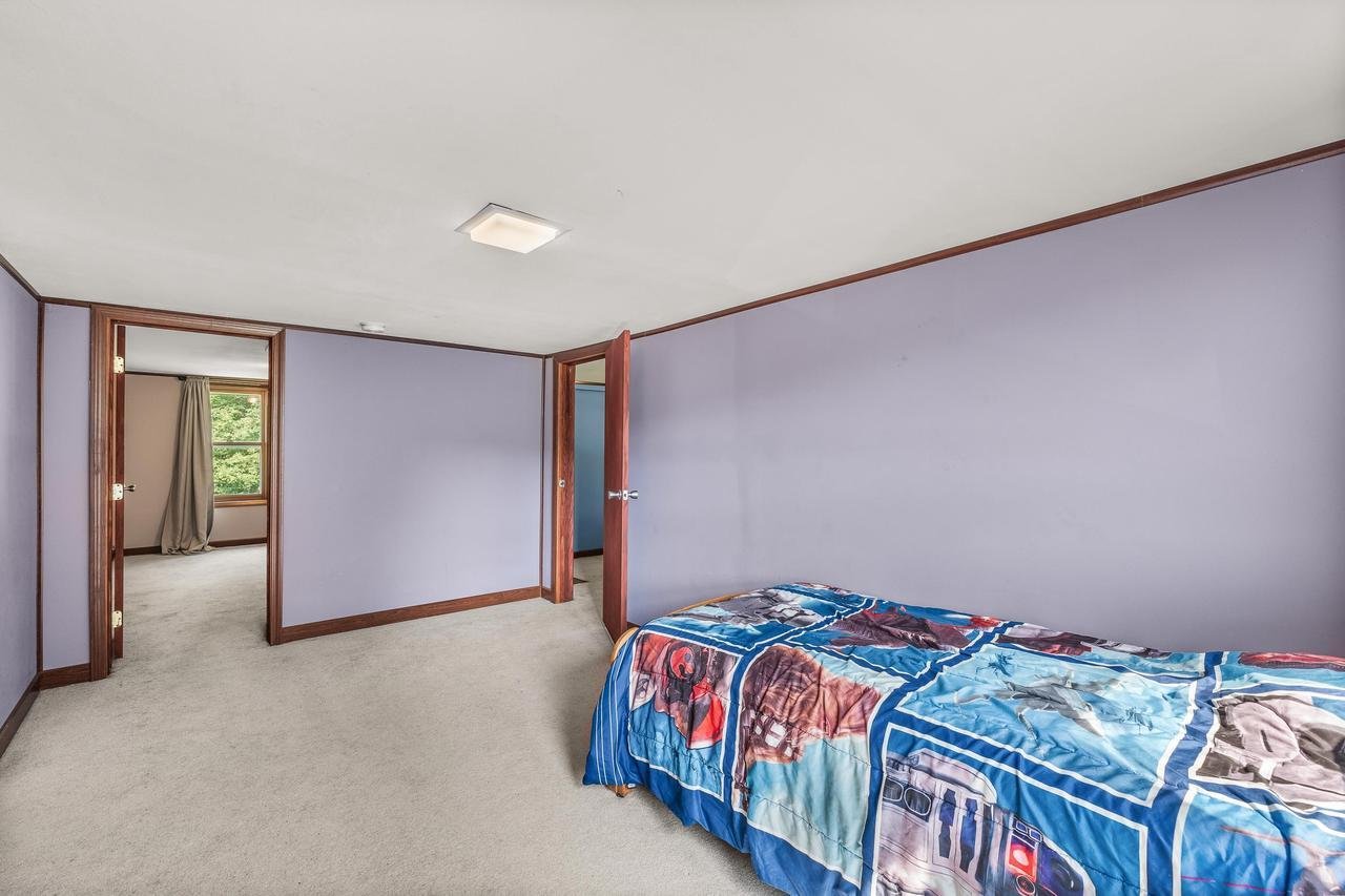 Small bedroom with purple walls, featuring a single bed covered with a colorful themed blanket, two open wooden doors leading to other rooms, and light-colored carpeting.
