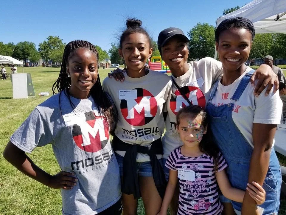 A group of five smiles outdoors on a sunny day at a community event. Four women in Mosaic Bible Fellowship T-shirts stand behind a young girl with colorful face paint, surrounded by grassy fields, tents, and a bounce house under a bright blue sky.
