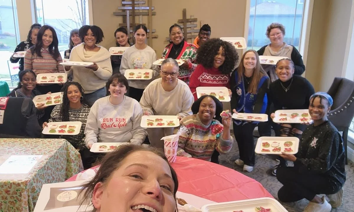 A group of women smiles and holds trays of decorated cookies during a festive gathering indoors. The setting includes Christmas-themed sweaters, tables with holiday tablecloths, and crosses on the wall in the background.