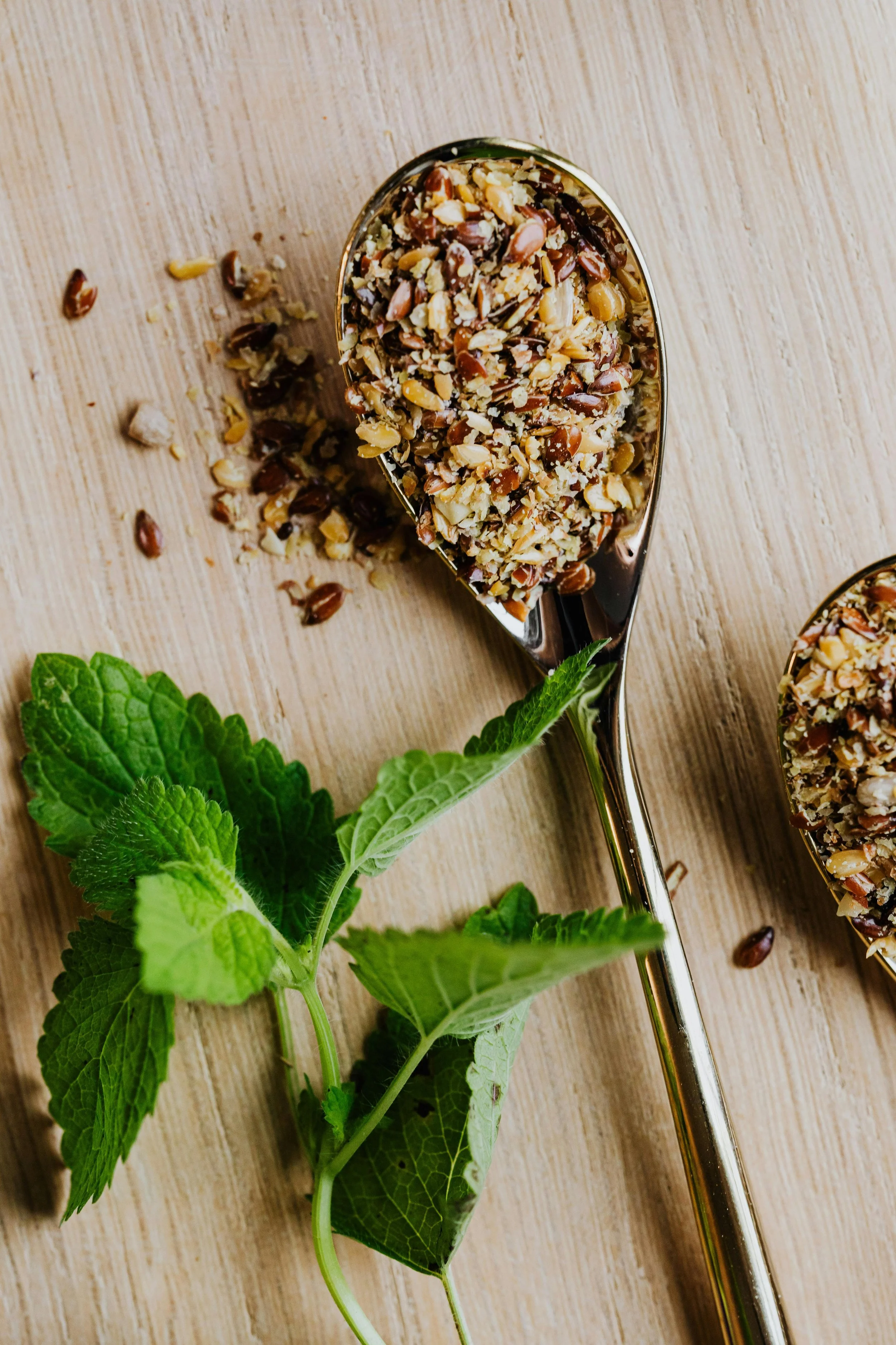 Two metal scoops filled with chopped nuts and seeds on a wooden surface, with fresh green mint leaves nearby.