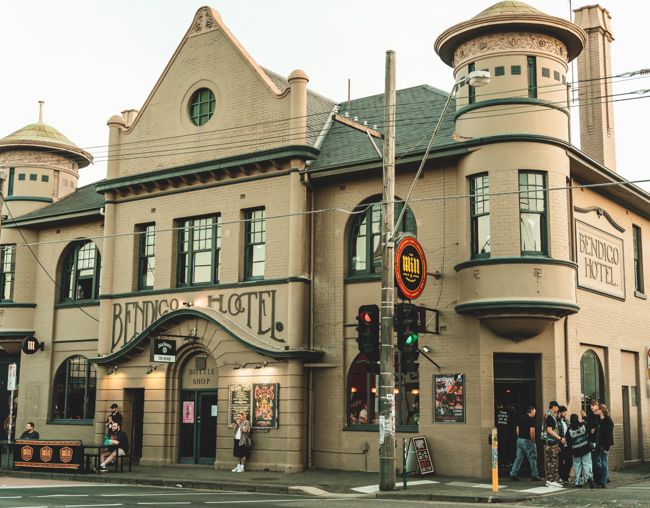 The Bendigo Hotel, a historic building with a beige brick facade and green trim, is located at street corner. There are people gathered outside, some sitting on benches and others waiting in line. Overhead, electric wires crisscross the street. The hotel has signage including a large sign reading "Bendigo Hotel" and a smaller sign for the bottle shop. The building features rounded turret-like towers and large windows.