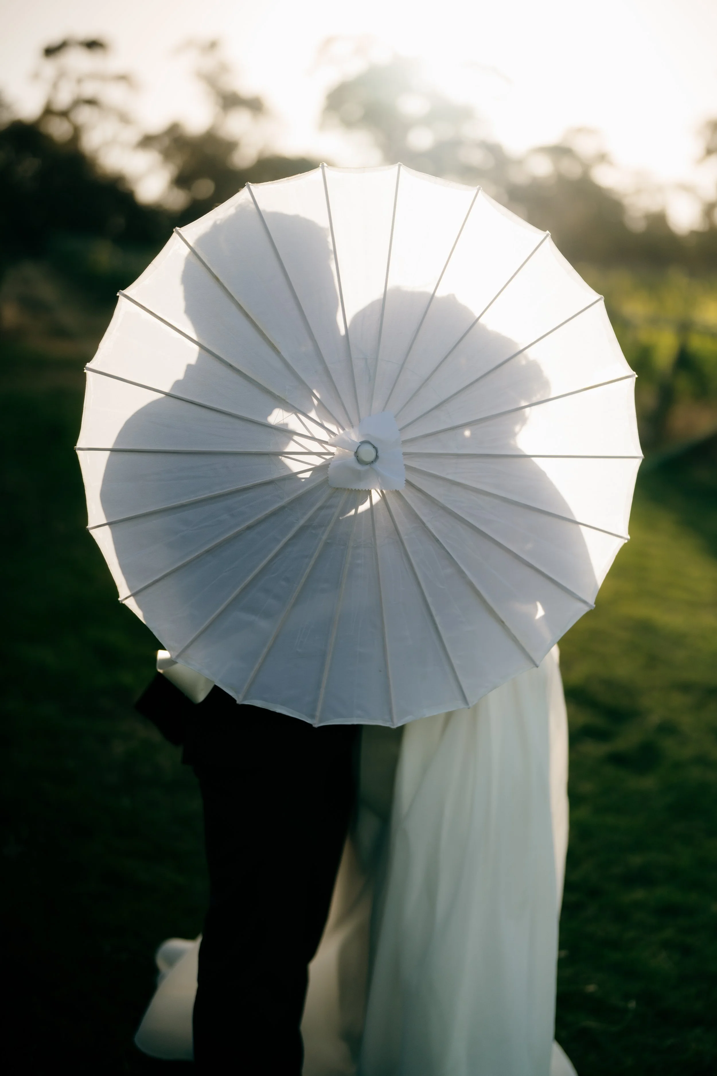 A couple in wedding attire standing outdoors, partially visible behind a large white umbrella, with sunlight in the background.