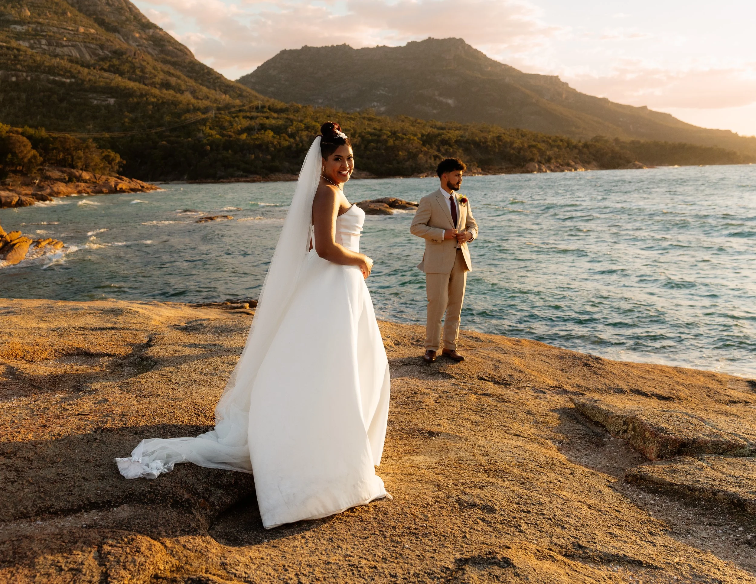 A bride in a white wedding dress and veil standing on rocks near the water with a groom in a tan suit behind her, during sunset with mountains in the background.