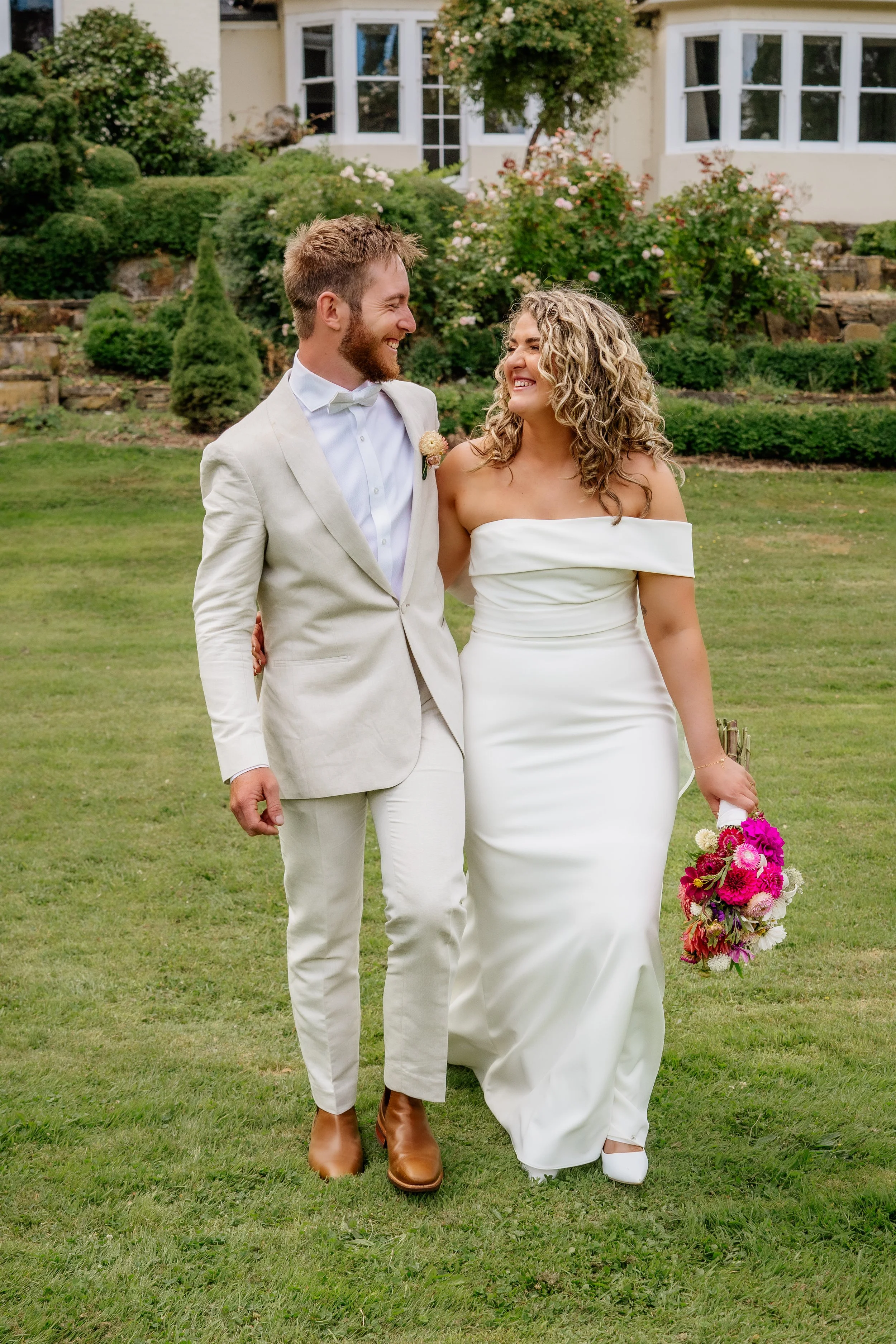 A happy couple, dressed in wedding attire, walking on a lush green lawn with a house and garden in the background.