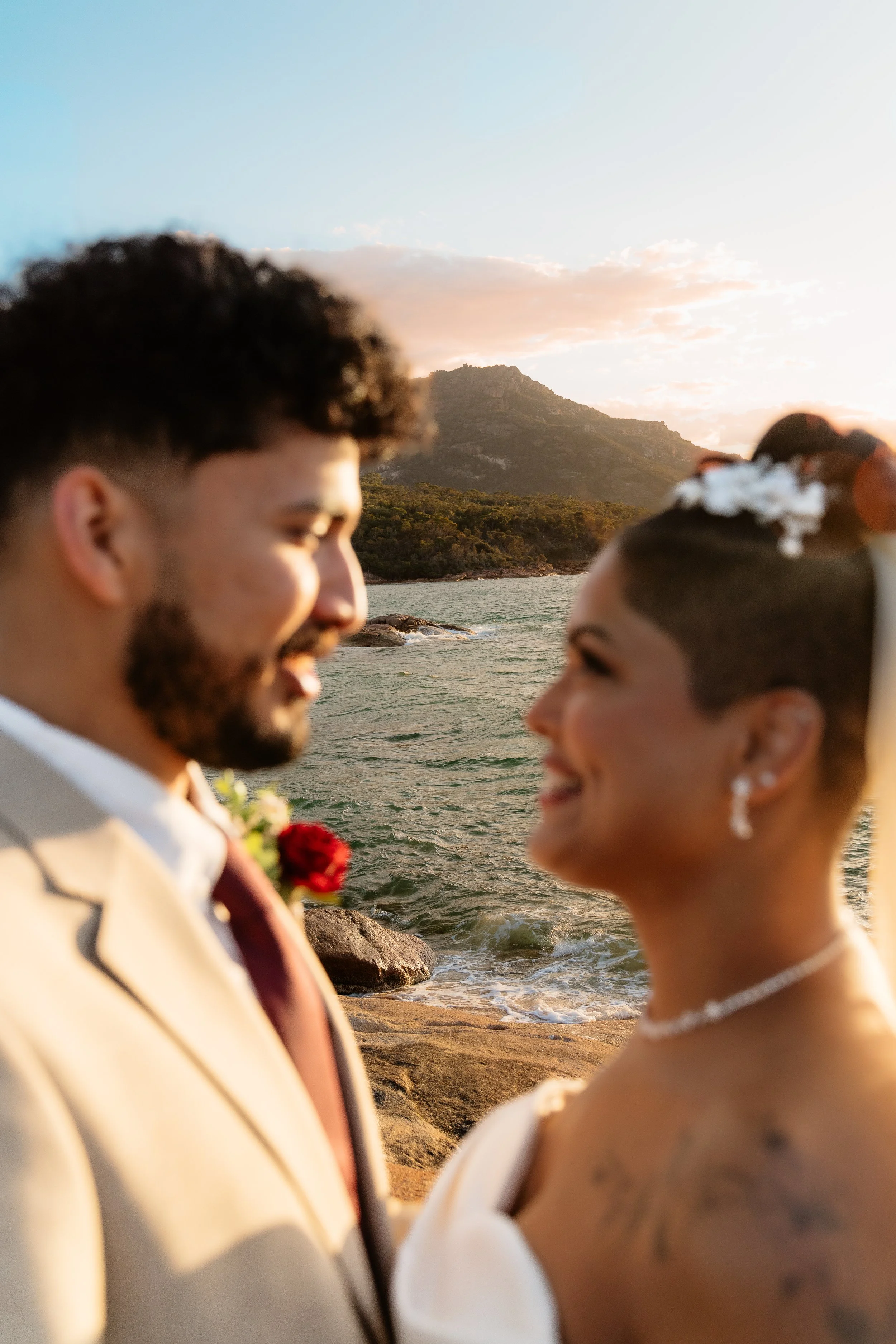 A bride and groom facing each other by a body of water at sunset, with mountains in the background.