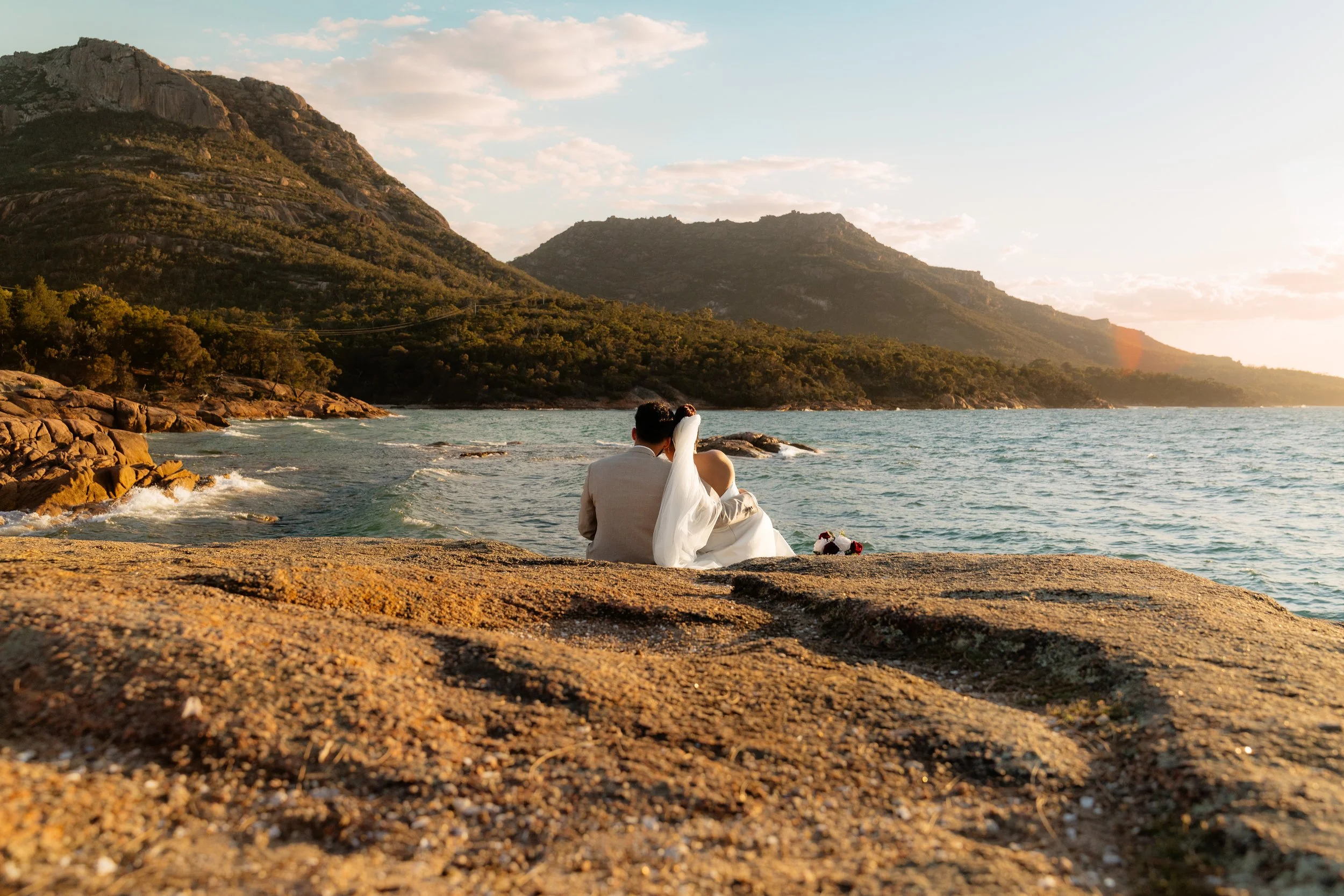 A bride and groom sitting together on a rocky shore by the ocean during sunset, with mountains in the background.