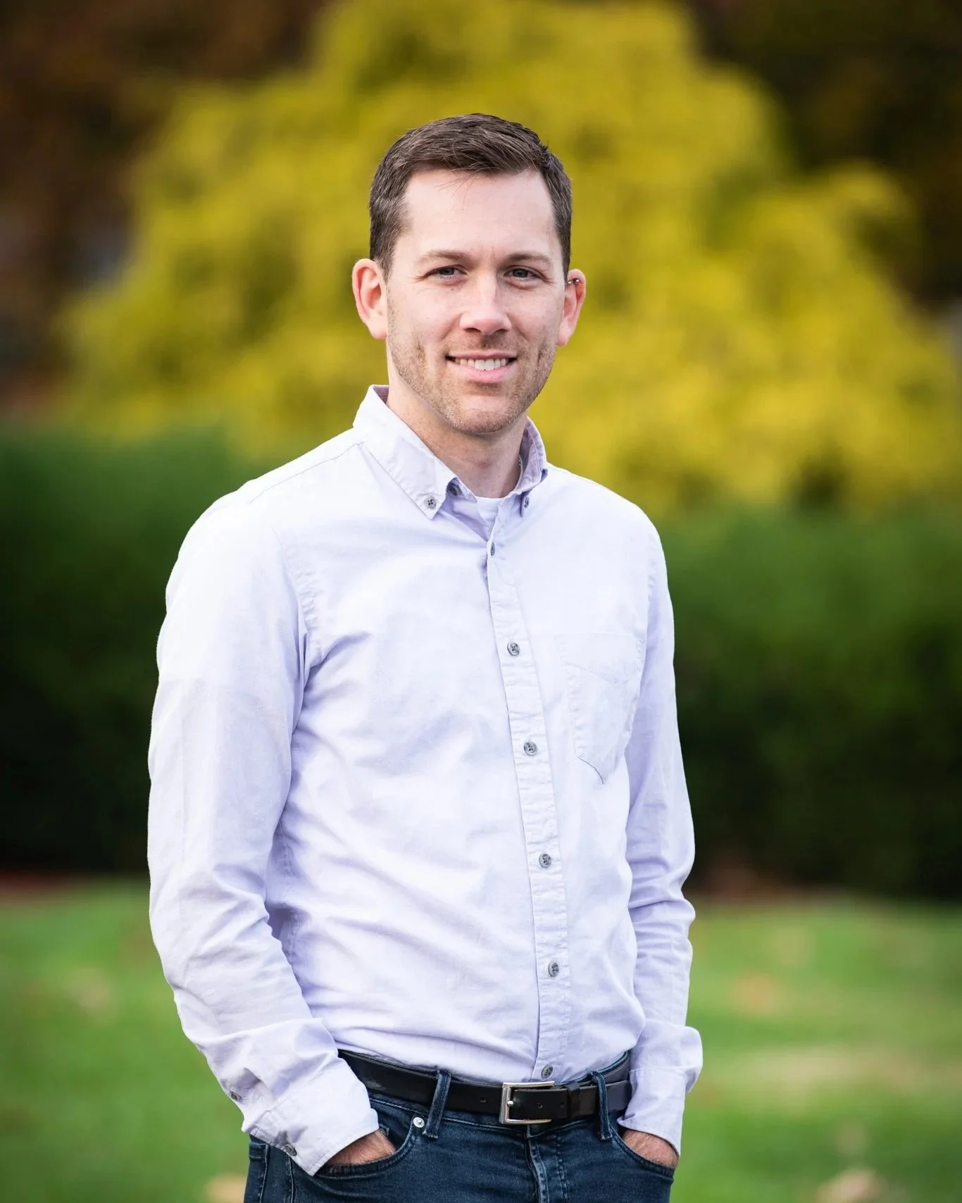 A young man with short brown hair and light skin, wearing a white button-up shirt and dark jeans, standing outdoors on a grassy area with green and yellow foliage in the background.