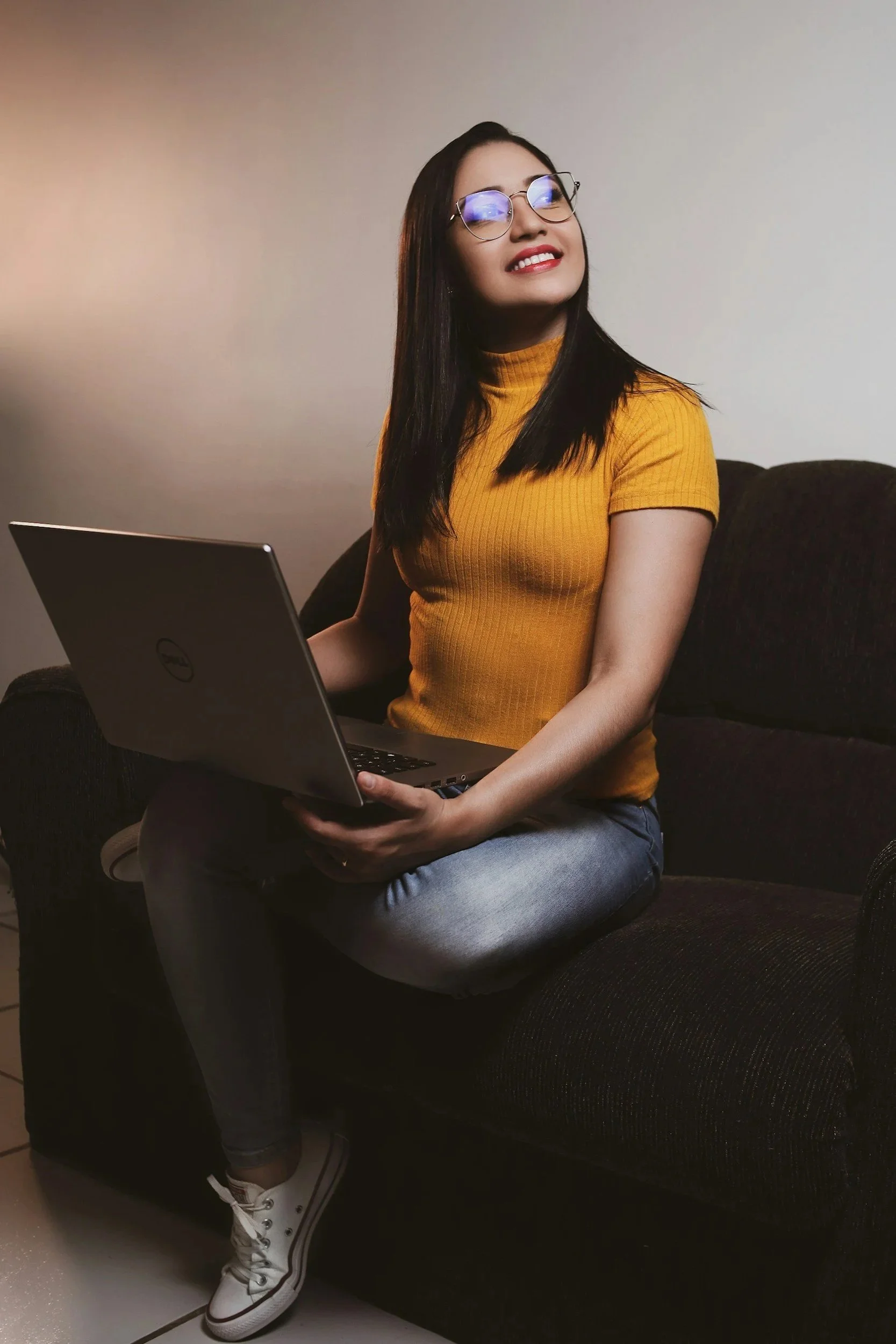 Woman sitting on a couch with a laptop open on her lap, smiling as she gazes off, representing the ease and accessibility of online therapy consultations in Virginia and Maryland